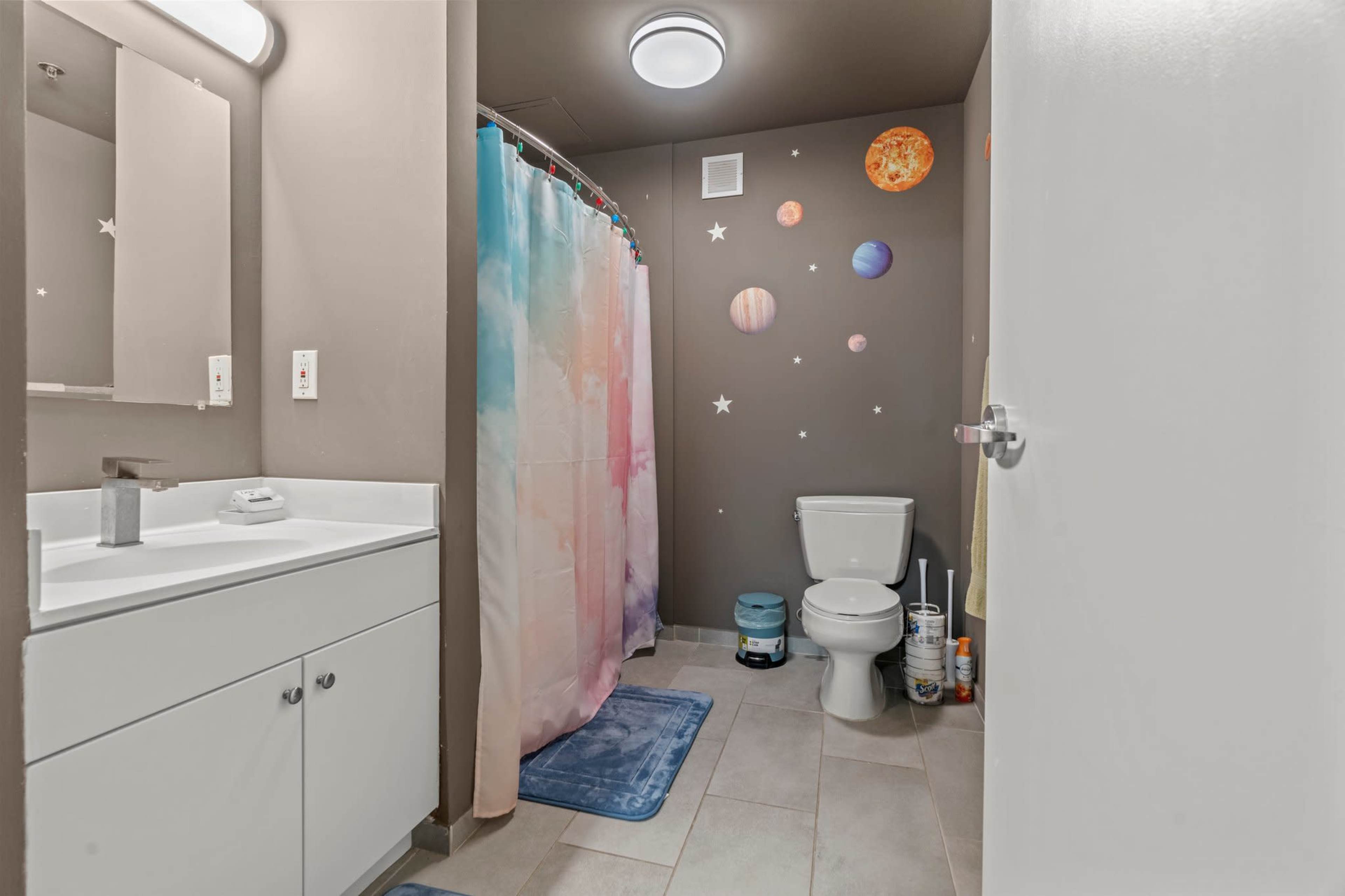 A bathroom features a colorful shower curtain with a galaxy design, a white toilet, and a sink with a mirror, all set against a wall painted with planets and stars.