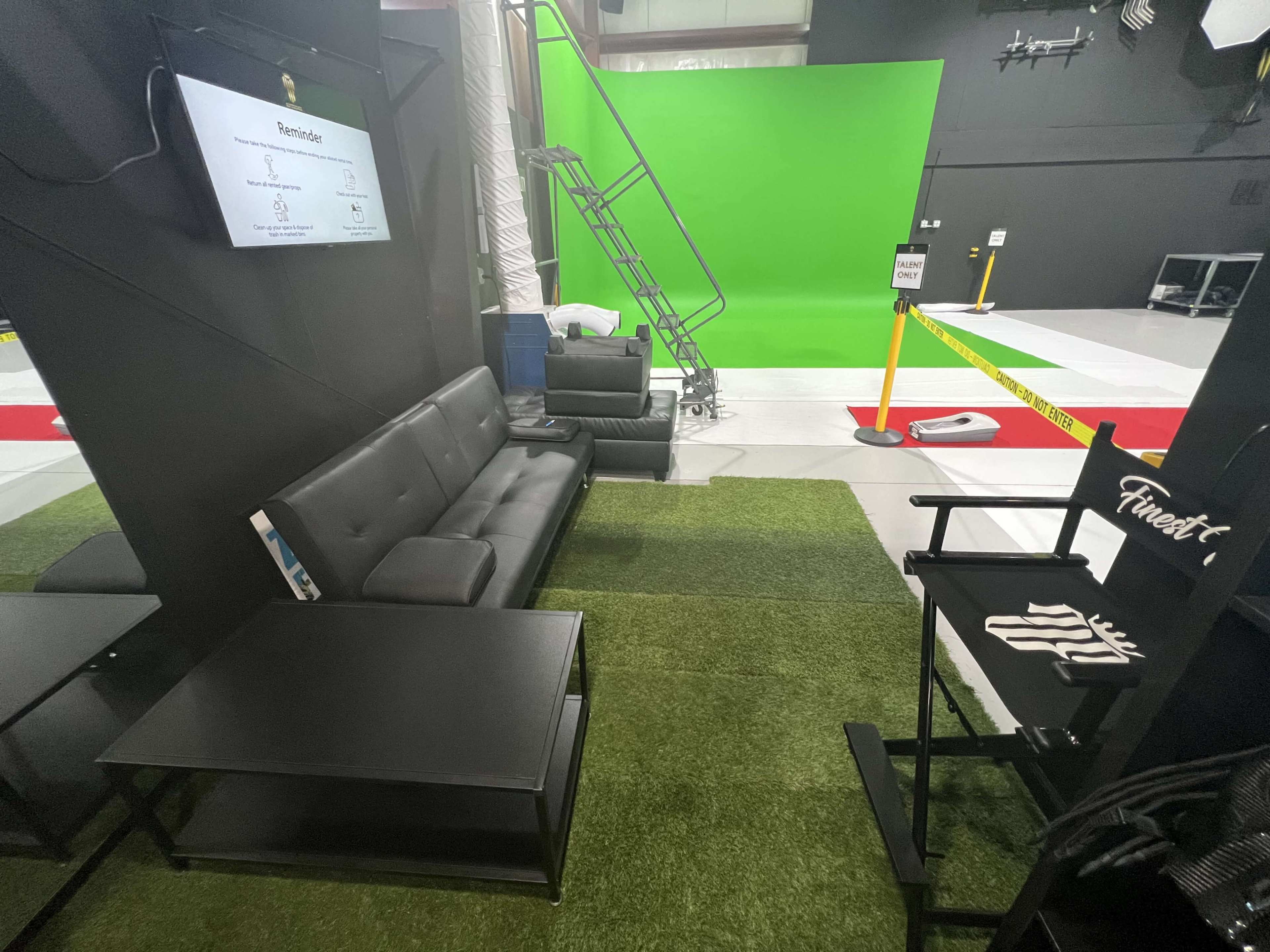The image shows a studio space with black sofas, tables, and a tall ladder leading to a green screen backdrop.