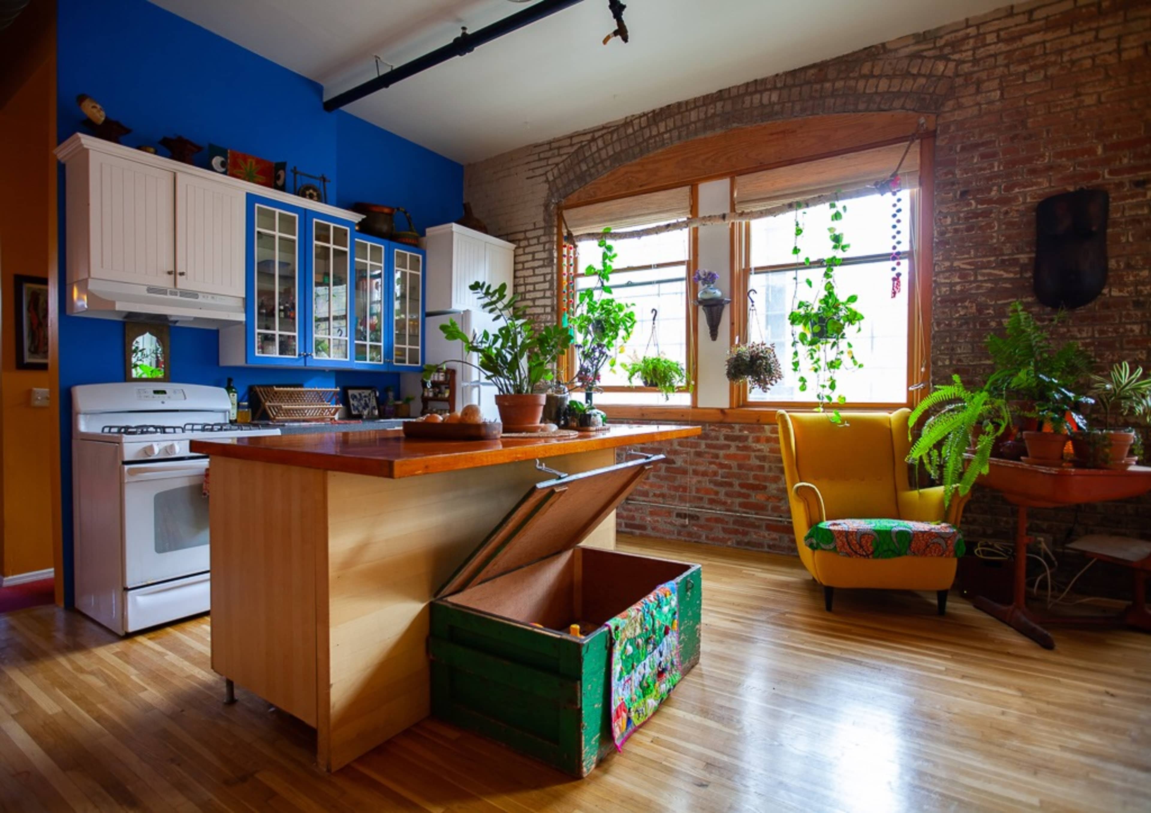 A cozy kitchen with a wooden island, a gas stove, and large windows adorned with various indoor plants, set against exposed brick walls.
