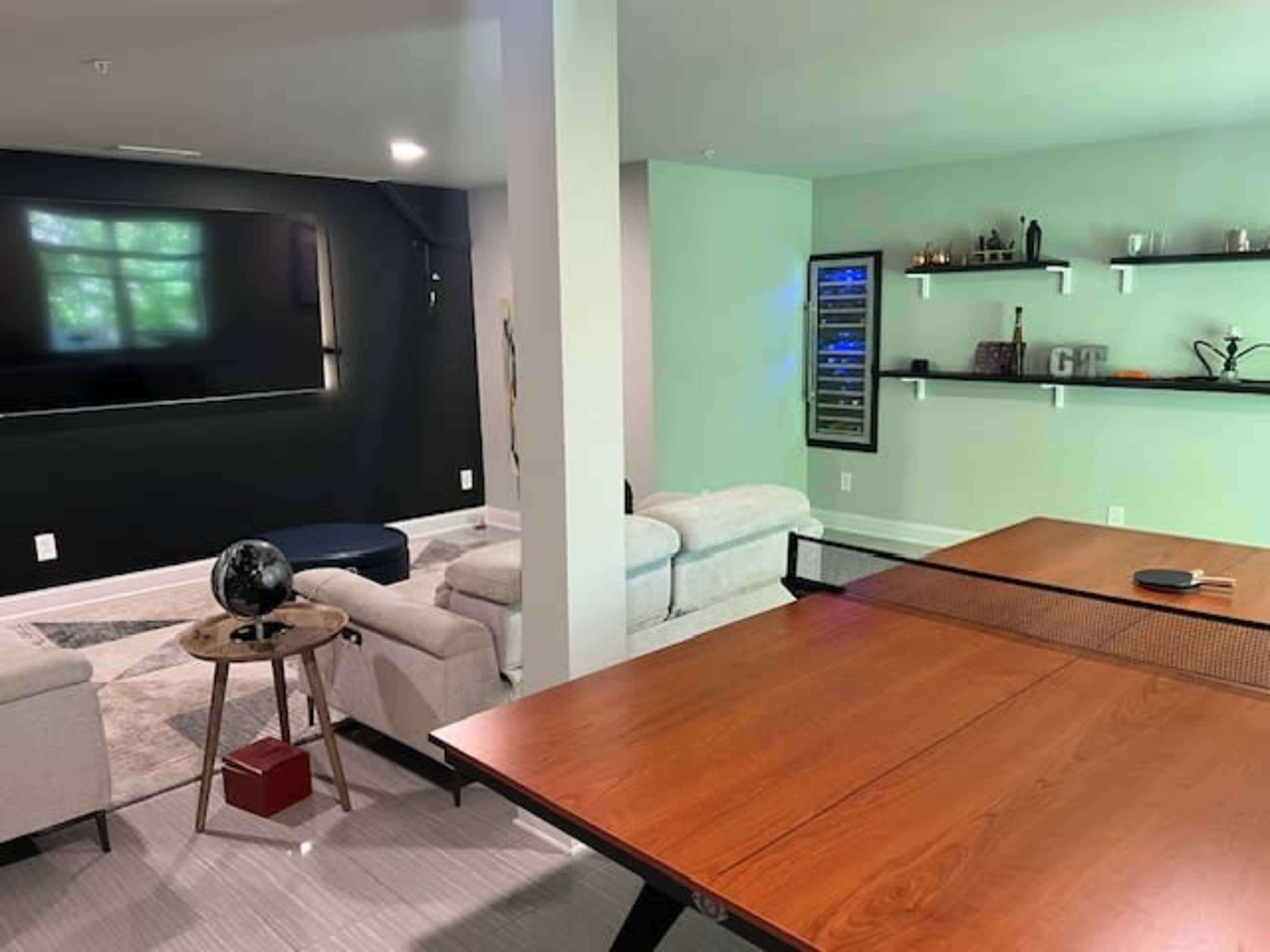 The image shows a modern basement living area featuring a large wall-mounted television, a comfortable seating arrangement, a ping pong table, and a beverage cooler on a shelf.