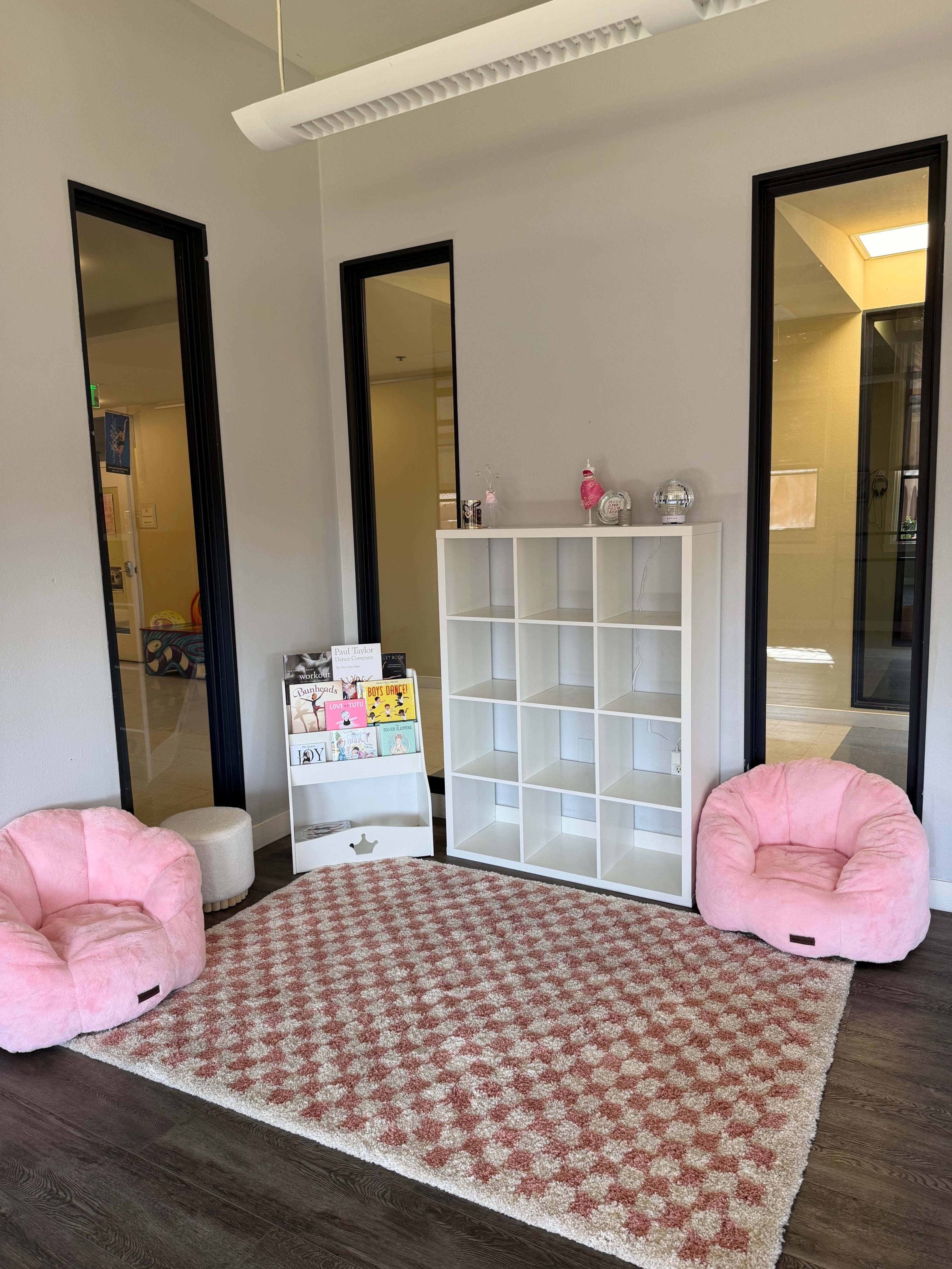 The image shows a small room with a pink and white patterned rug, two pink beanbag chairs, a shelving unit, and a display stand with children's artwork.