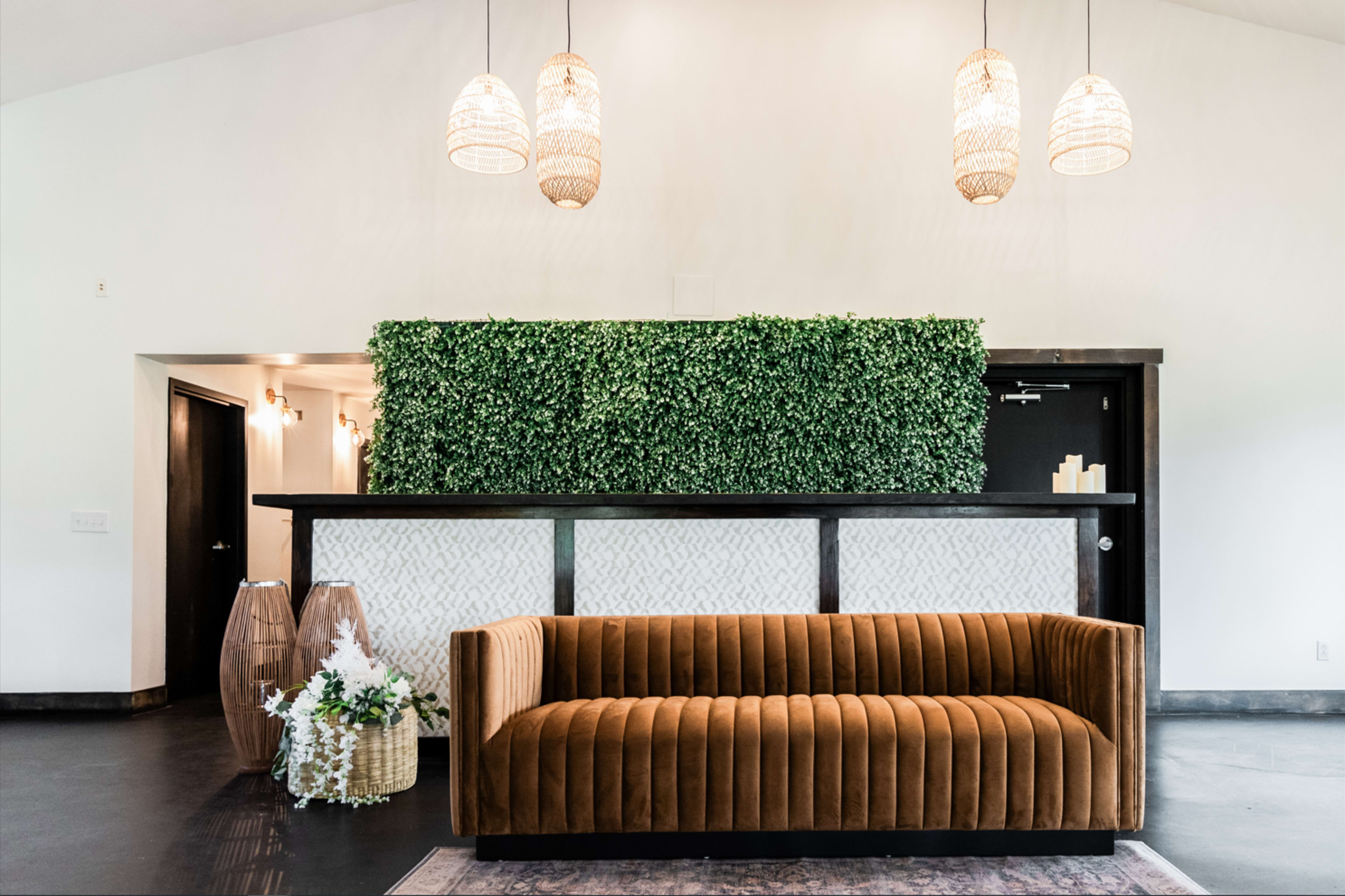 A modern reception area featuring a brown velvet sofa, a green leafy panel behind a wooden counter, and pendant lighting.