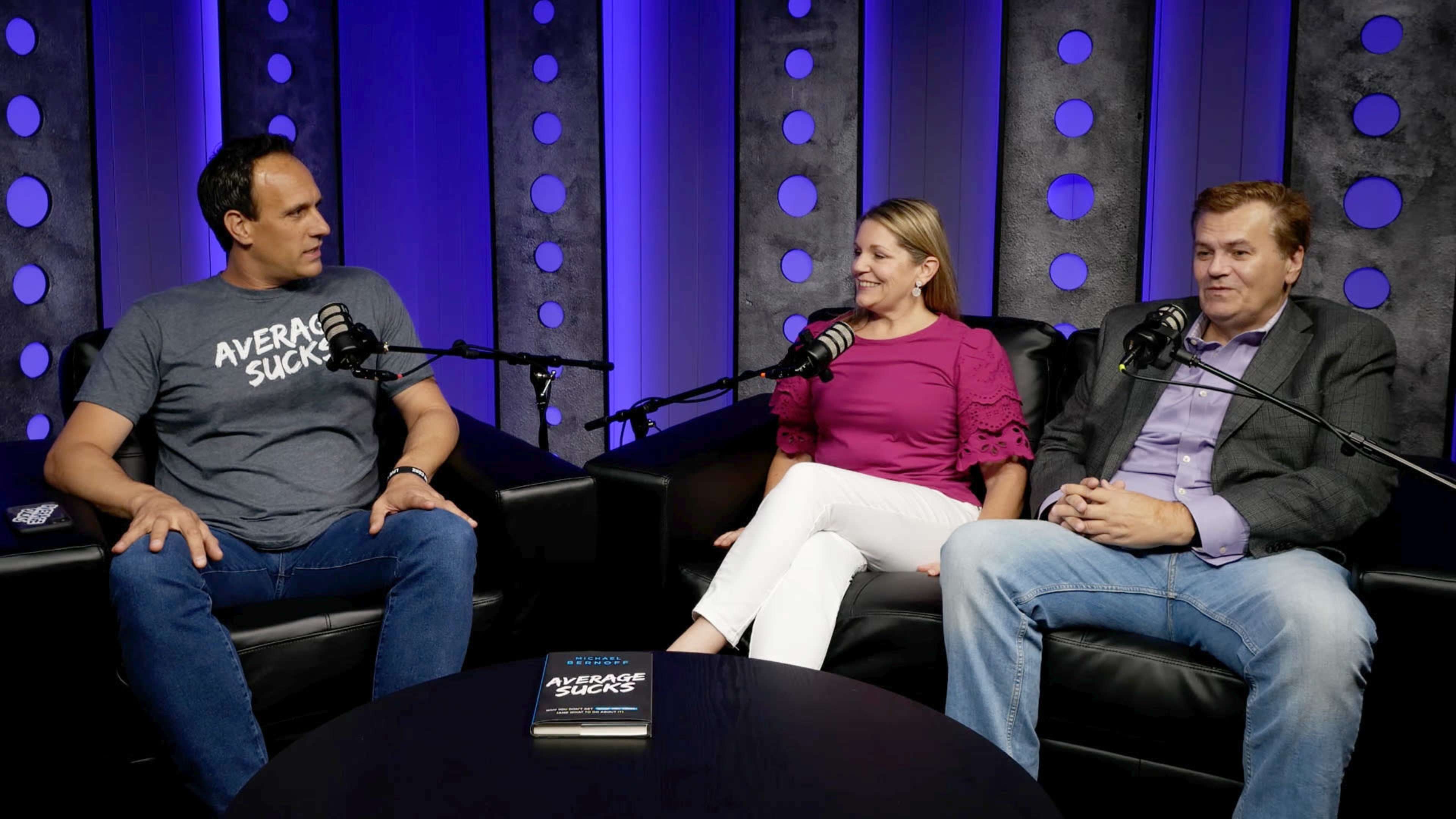 Three people are seated on a black couch in front of microphones, discussing a book titled "Average Sucks," with a blue background featuring circular patterns.