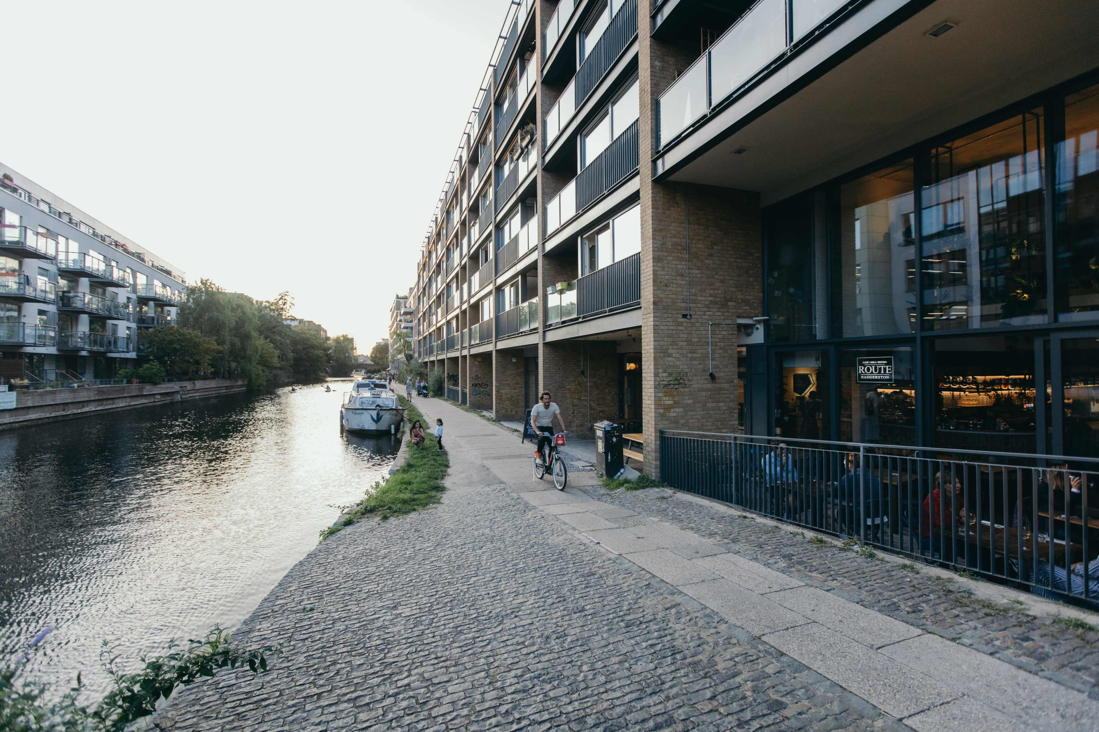 A cyclist rides along a cobblestone path beside a canal, with modern buildings and a boat moored nearby.