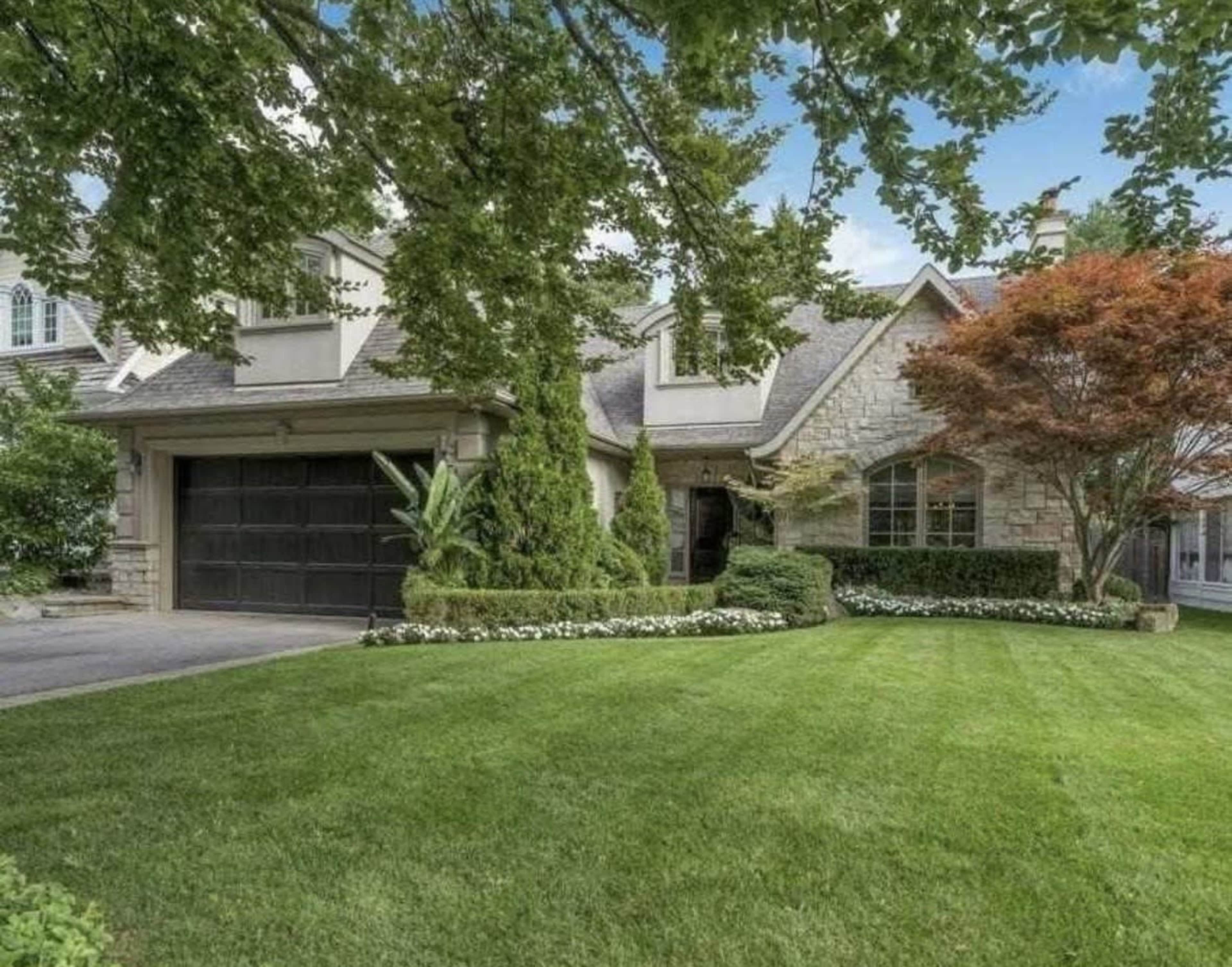 The image shows a well-maintained house with a stone exterior, a manicured lawn, and neatly trimmed shrubs and trees.