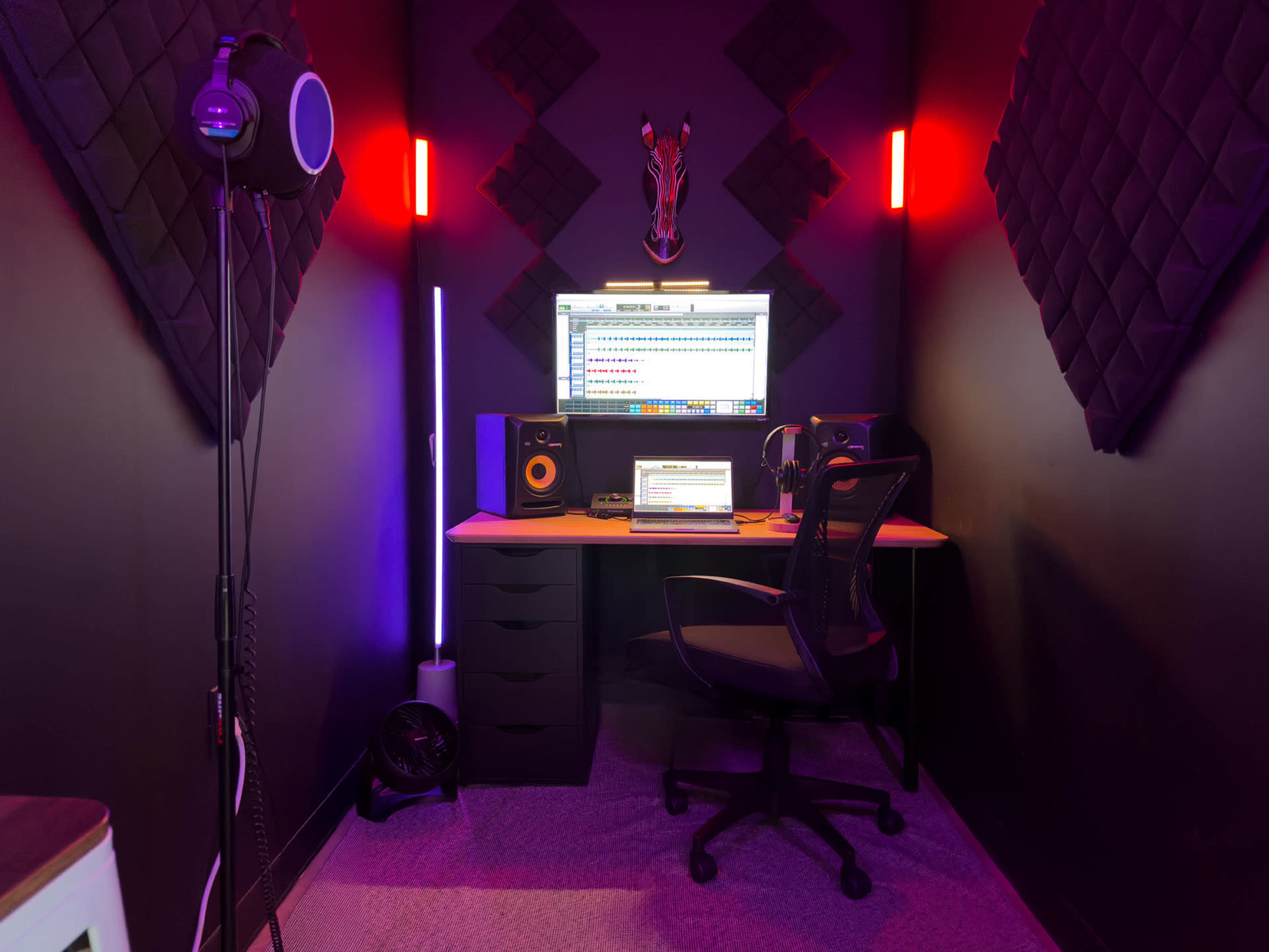The image shows a small, dark recording studio featuring a desk with a computer, speakers, and a headset, illuminated by colorful LED lights and decorated with sound-absorbing panels.