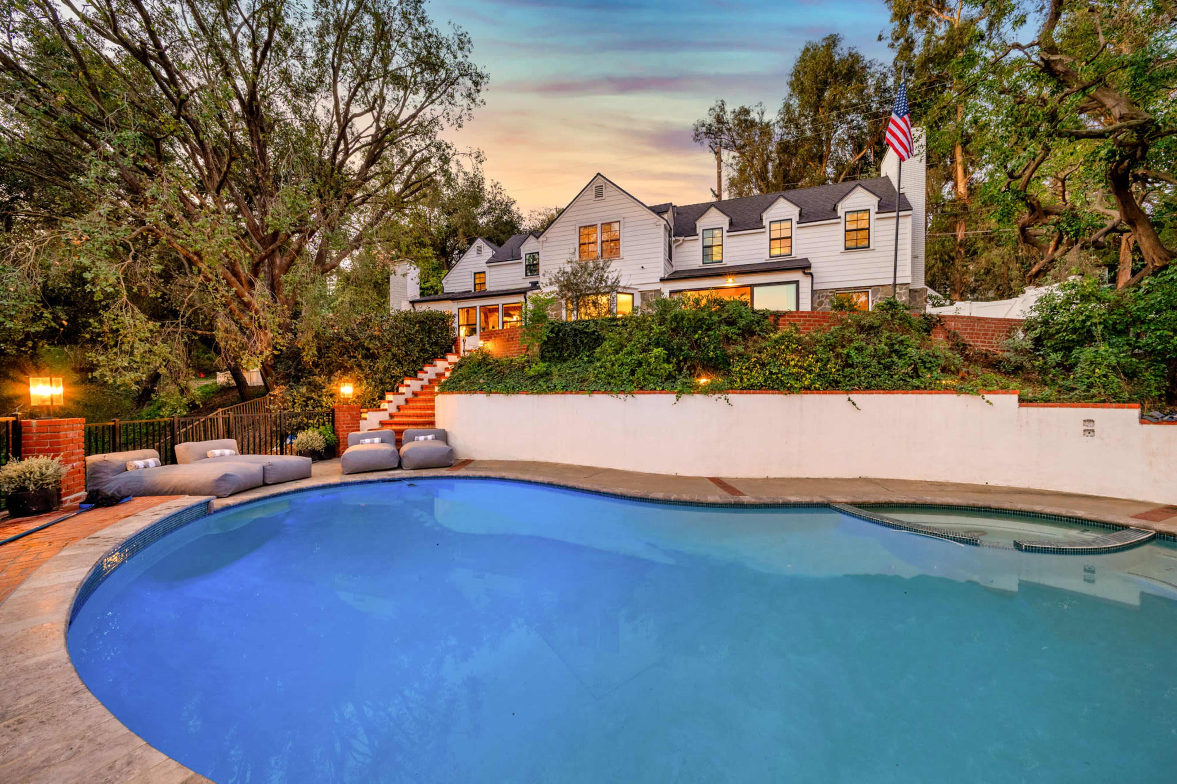 A large, blue swimming pool is surrounded by landscaped greenery and a two-story house with a flag in the background.