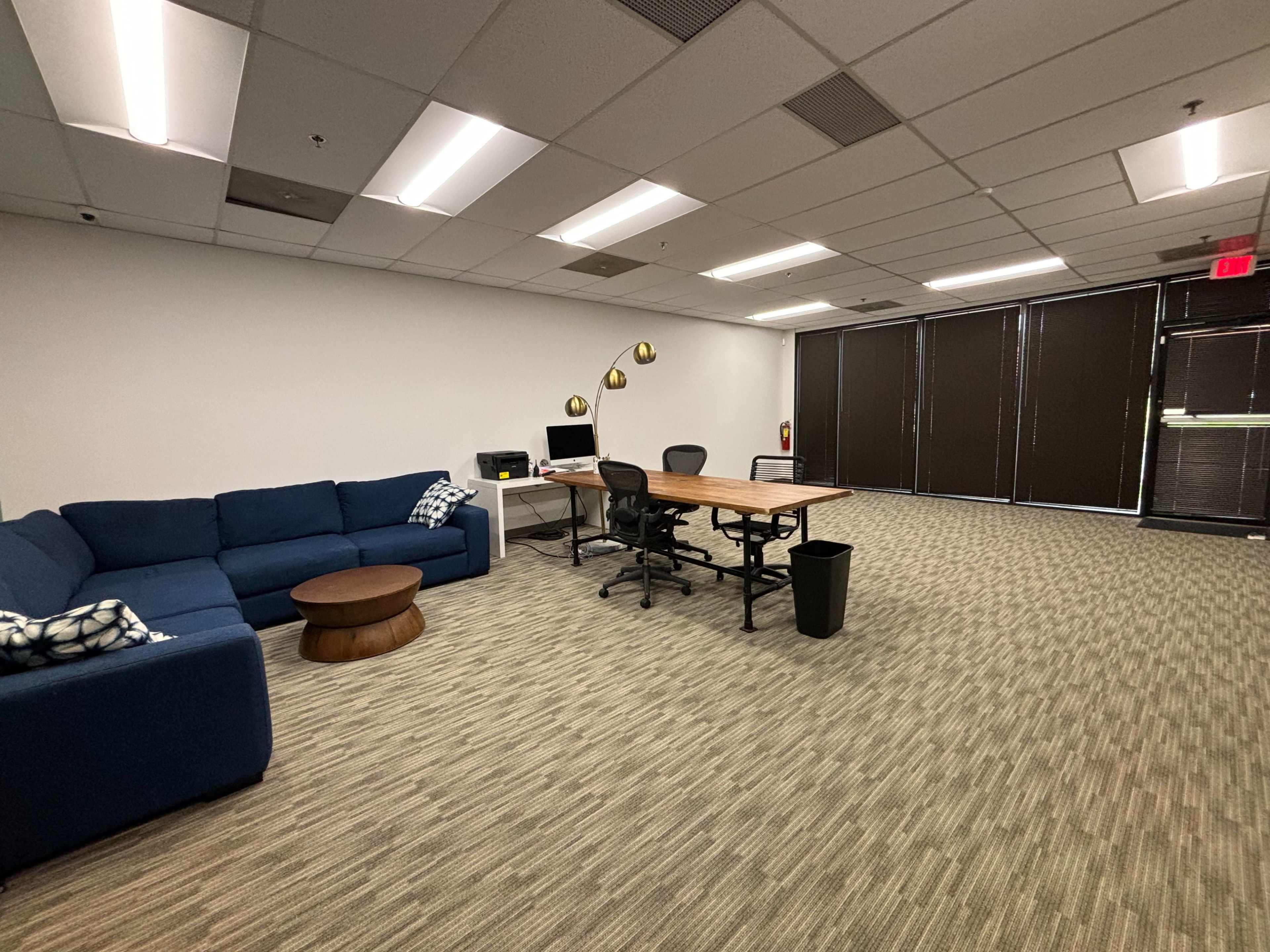 A modern office space with a blue sofa, wooden coffee table, a desk with chairs, and large windows covered by blinds.