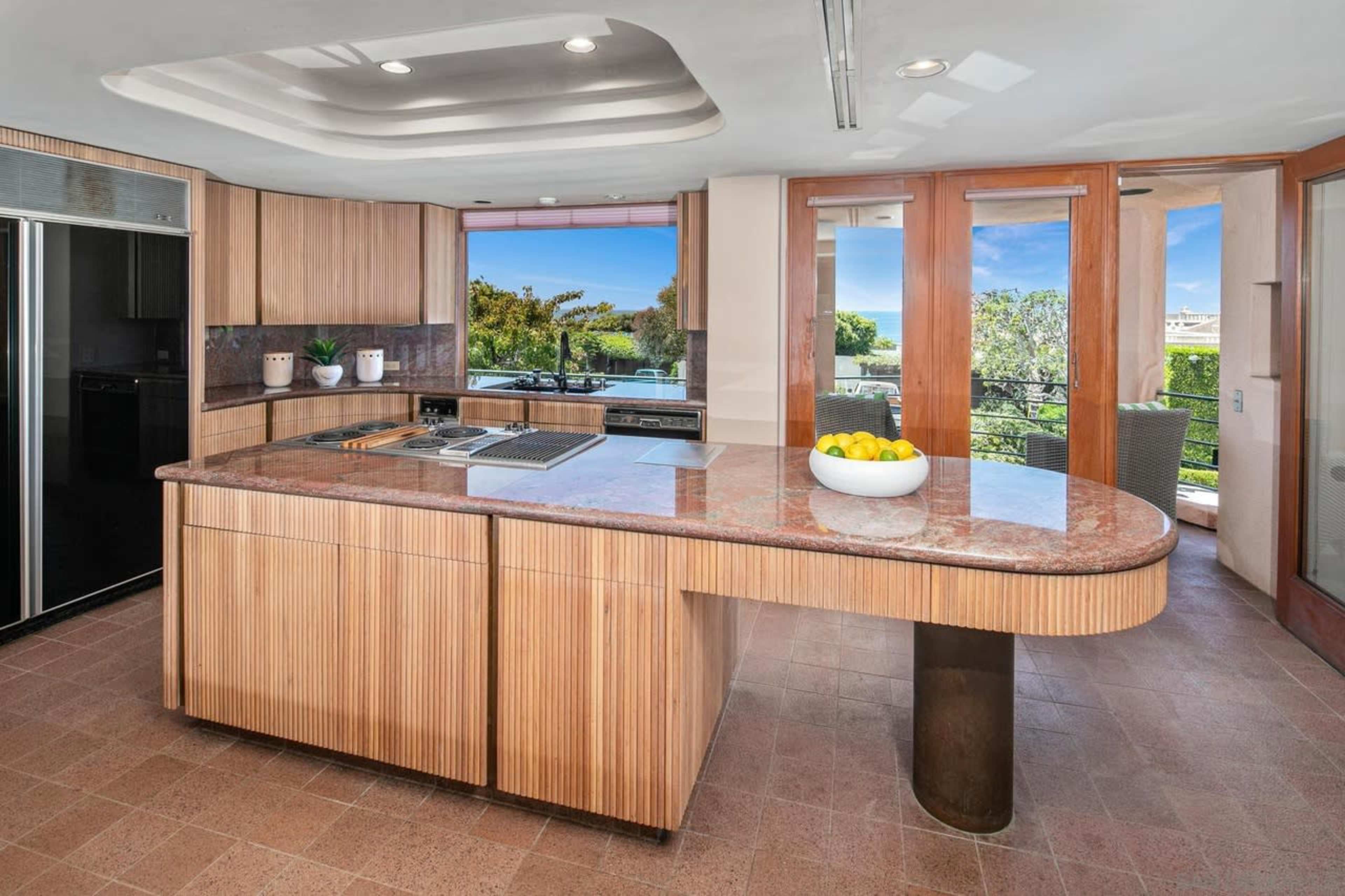 The image shows a modern kitchen with a large curved island, stainless steel appliances, and large windows overlooking an outdoor garden space.