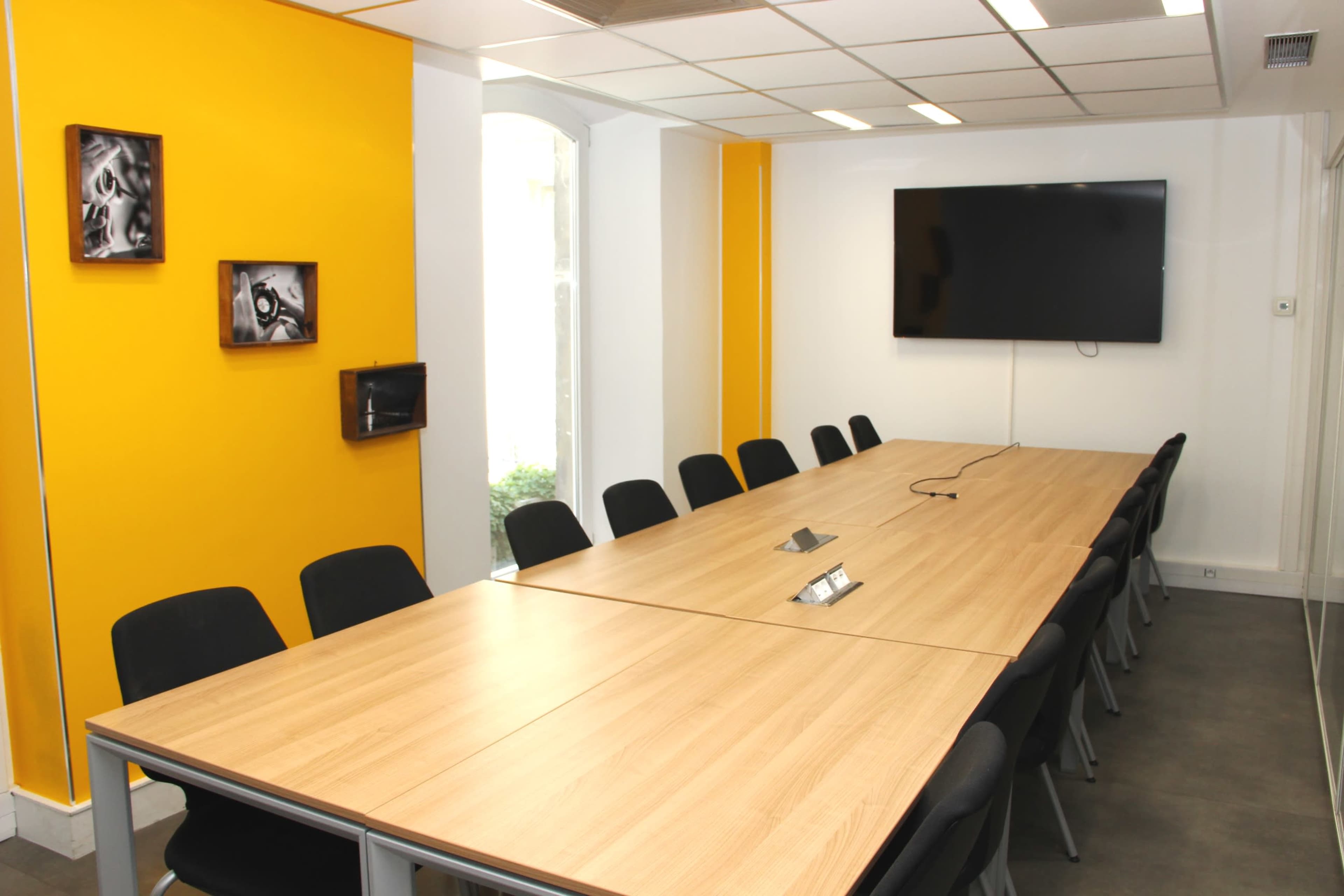 A meeting room with a long wooden table surrounded by black chairs, yellow accent walls, and a wall-mounted television.