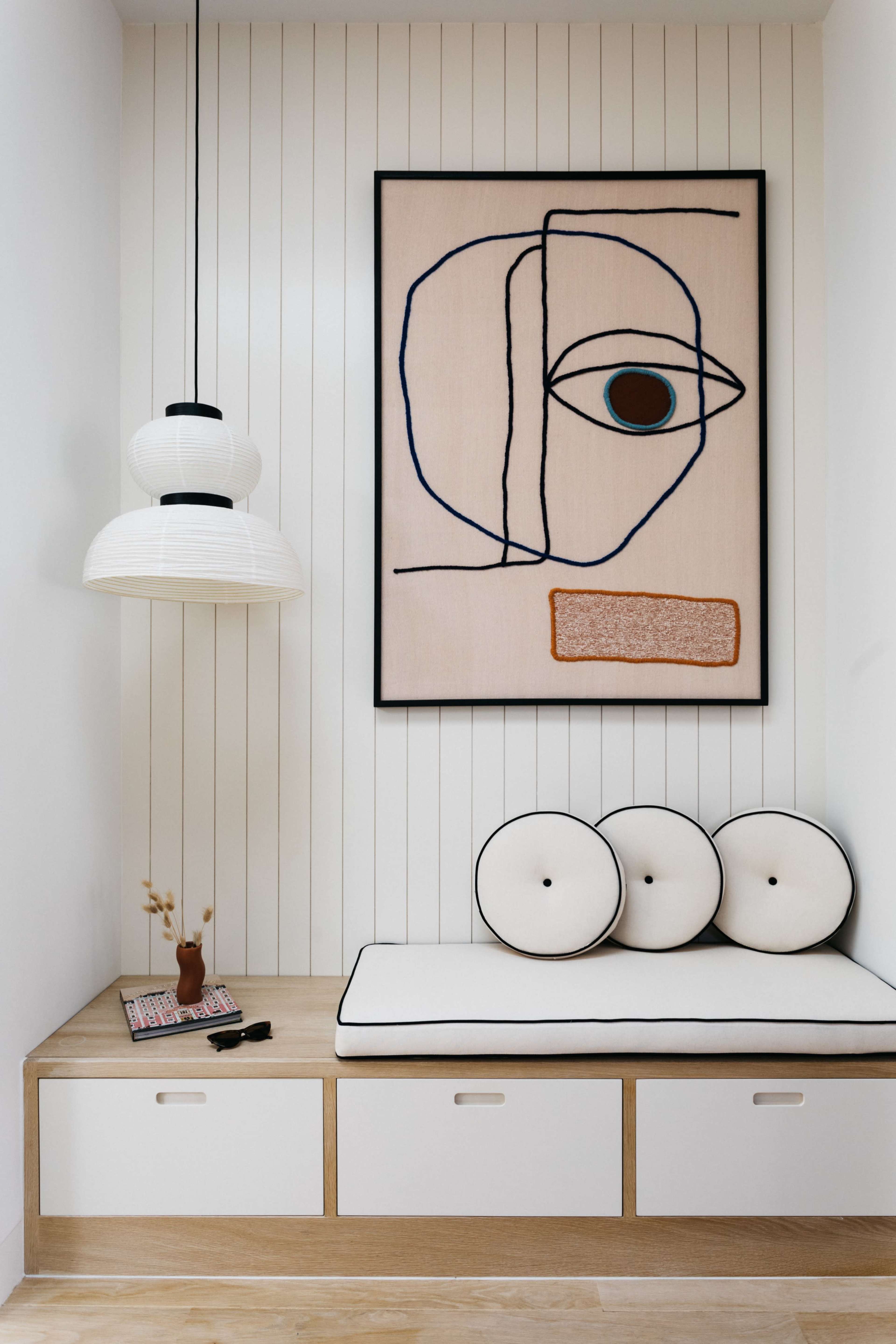 The image shows a modern nook featuring a padded bench with round cushions, a framed abstract artwork on the wall, and a pendant light hanging above.