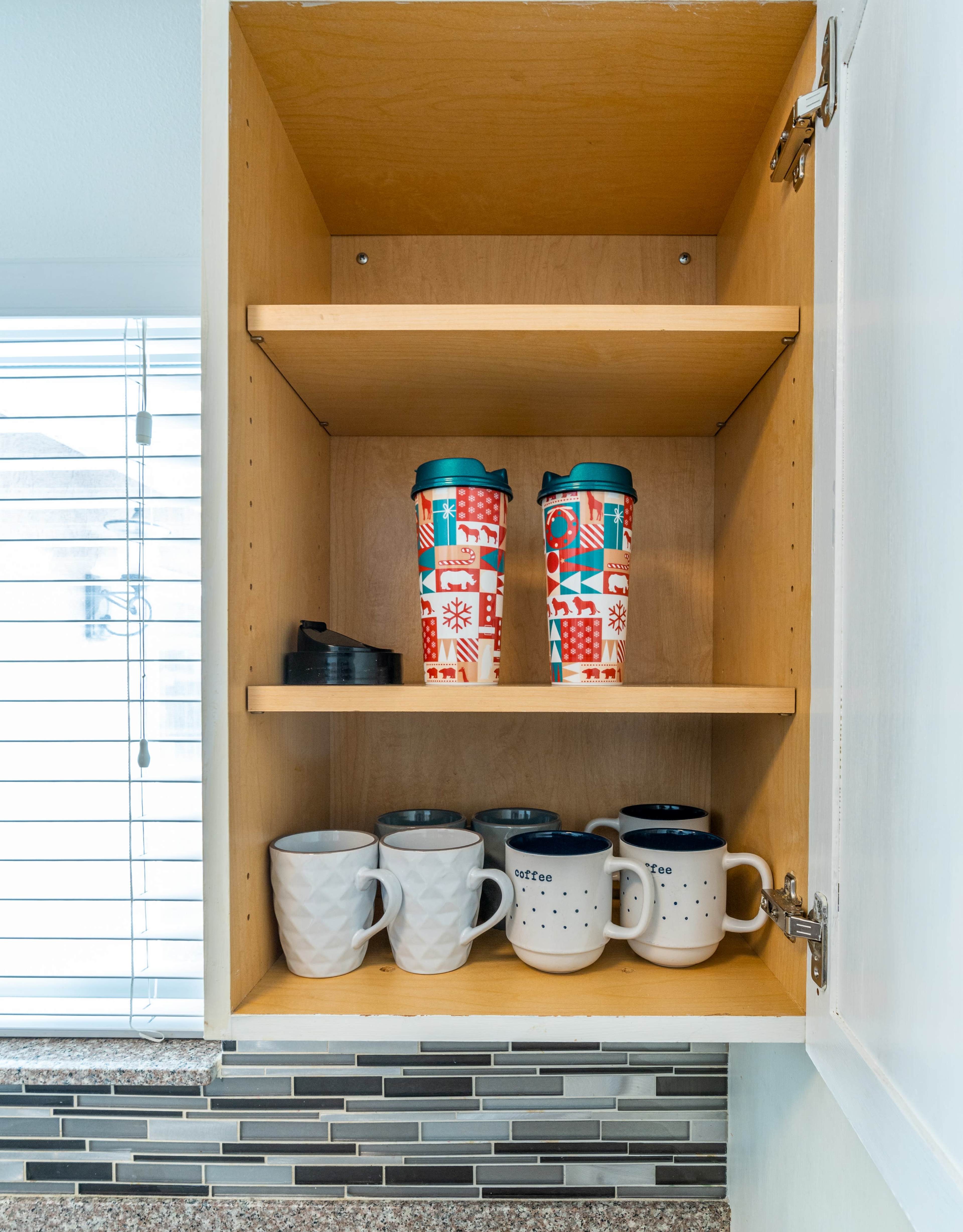 A kitchen cabinet contains mugs and travel tumblers organized on three shelves.