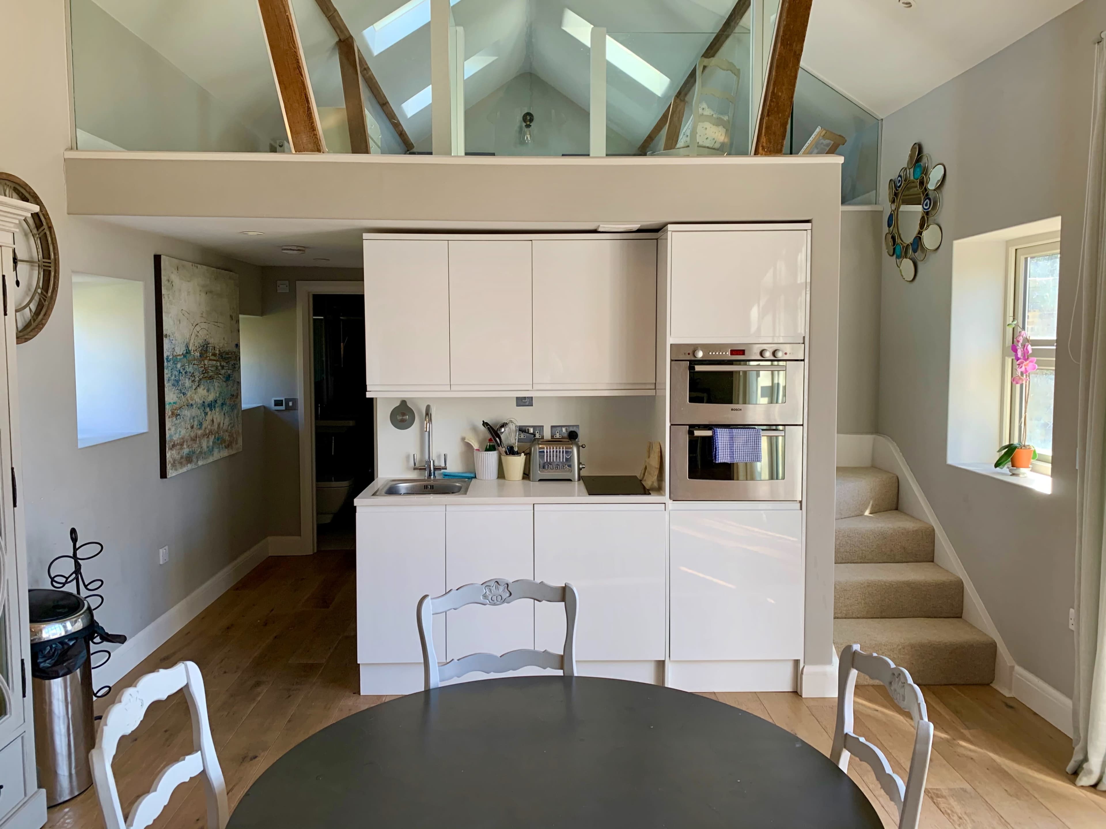 The image shows a modern kitchen with white cabinetry, a built-in oven, and a small dining table in front of it, alongside a staircase leading to an upper level.