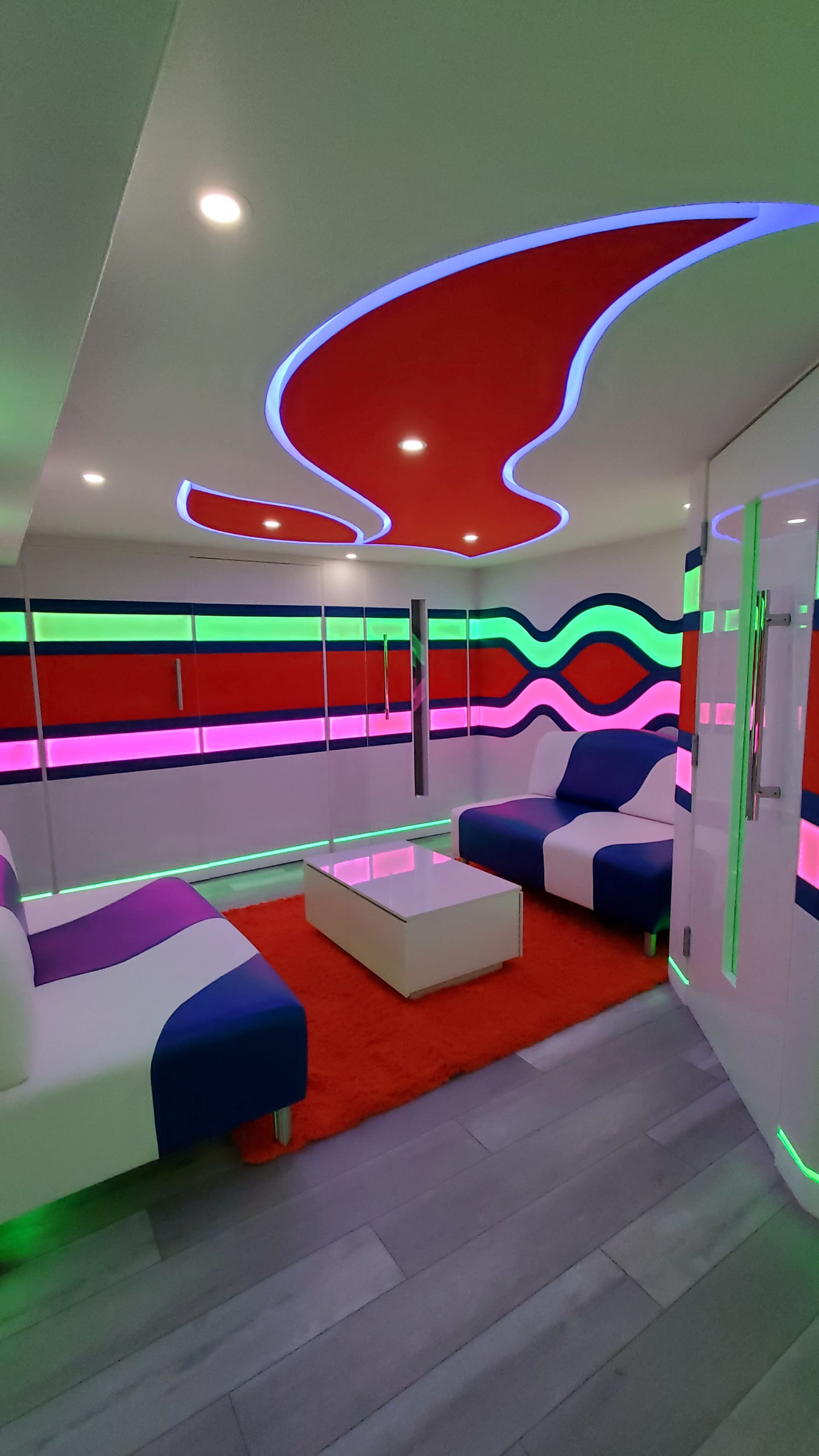 The room features bold, curving neon lights along the walls and ceiling, with modern furniture in a colorful design, including a white sofa with blue and purple accents on a vibrant red rug.