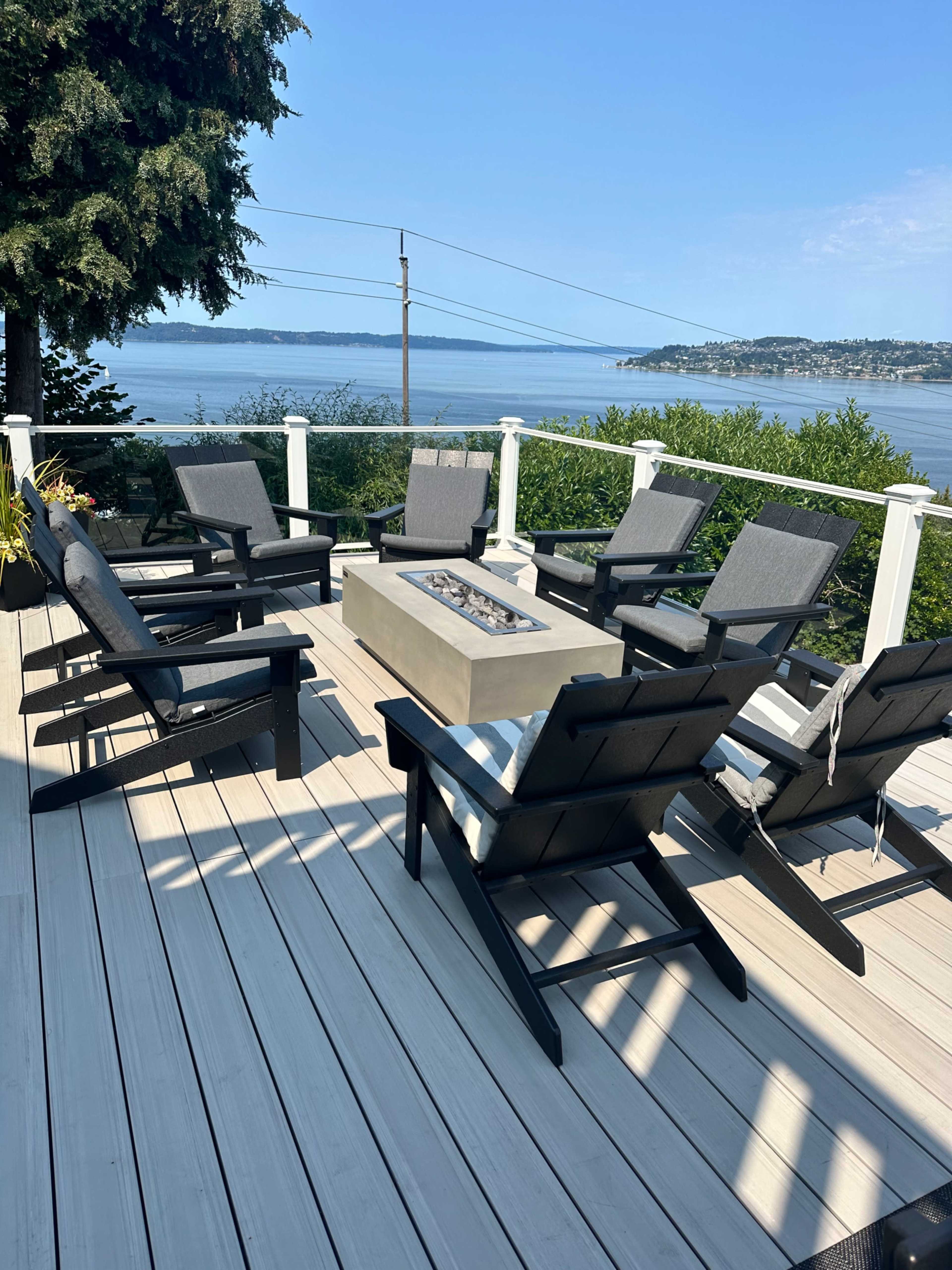 A patio area features six black lounge chairs arranged around a fire pit, overlooking a body of water and distant shoreline.