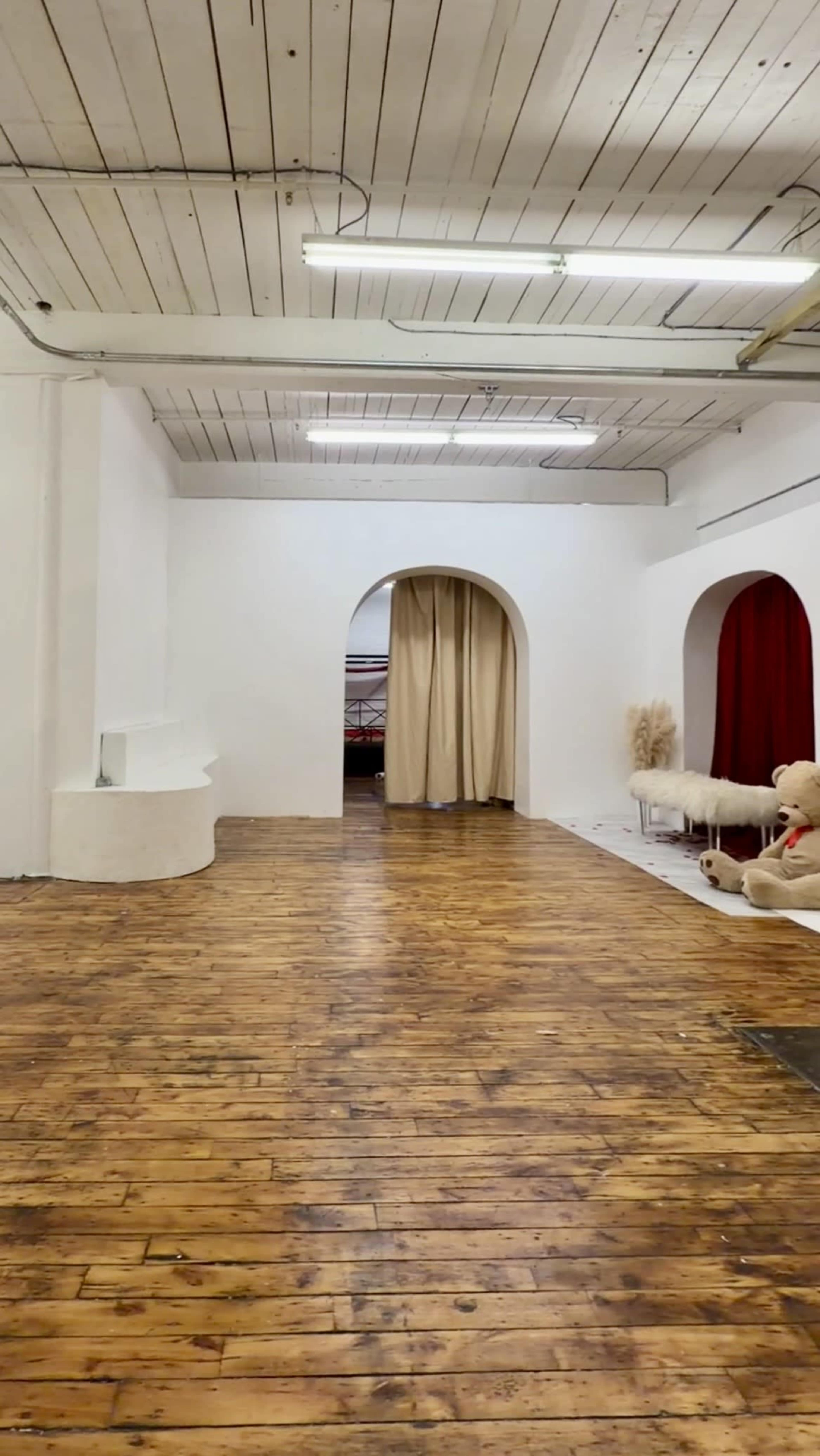 The image shows a spacious, empty room with wooden floors, arched doorways, and a curtain separating different areas.