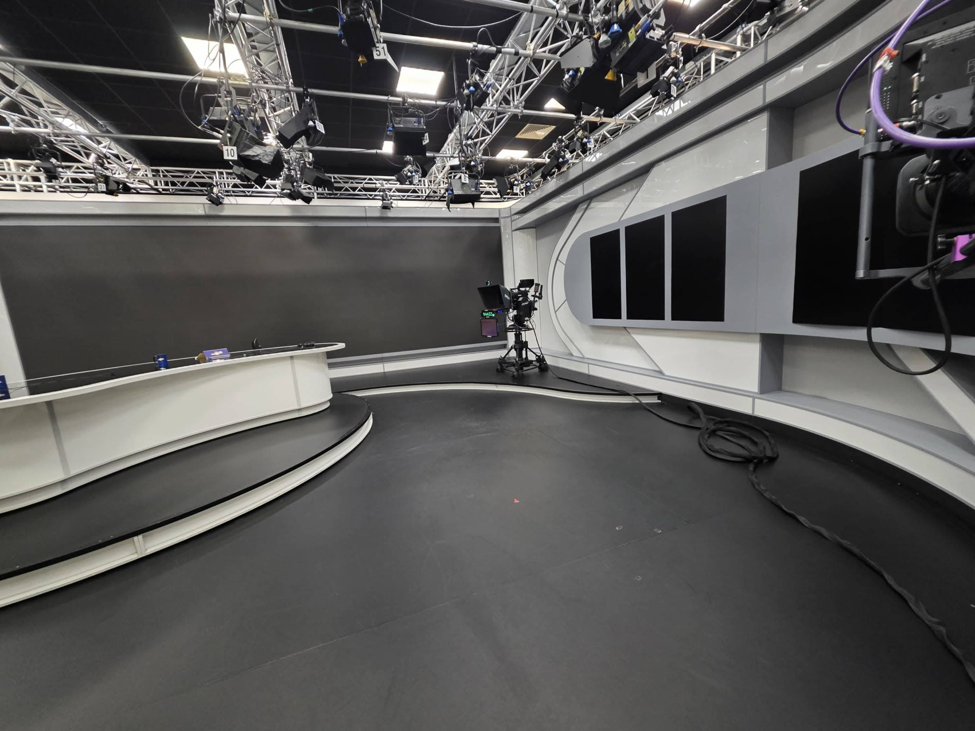 The image shows a modern television studio with a curved desk, camera equipment, and multiple overhead lights.