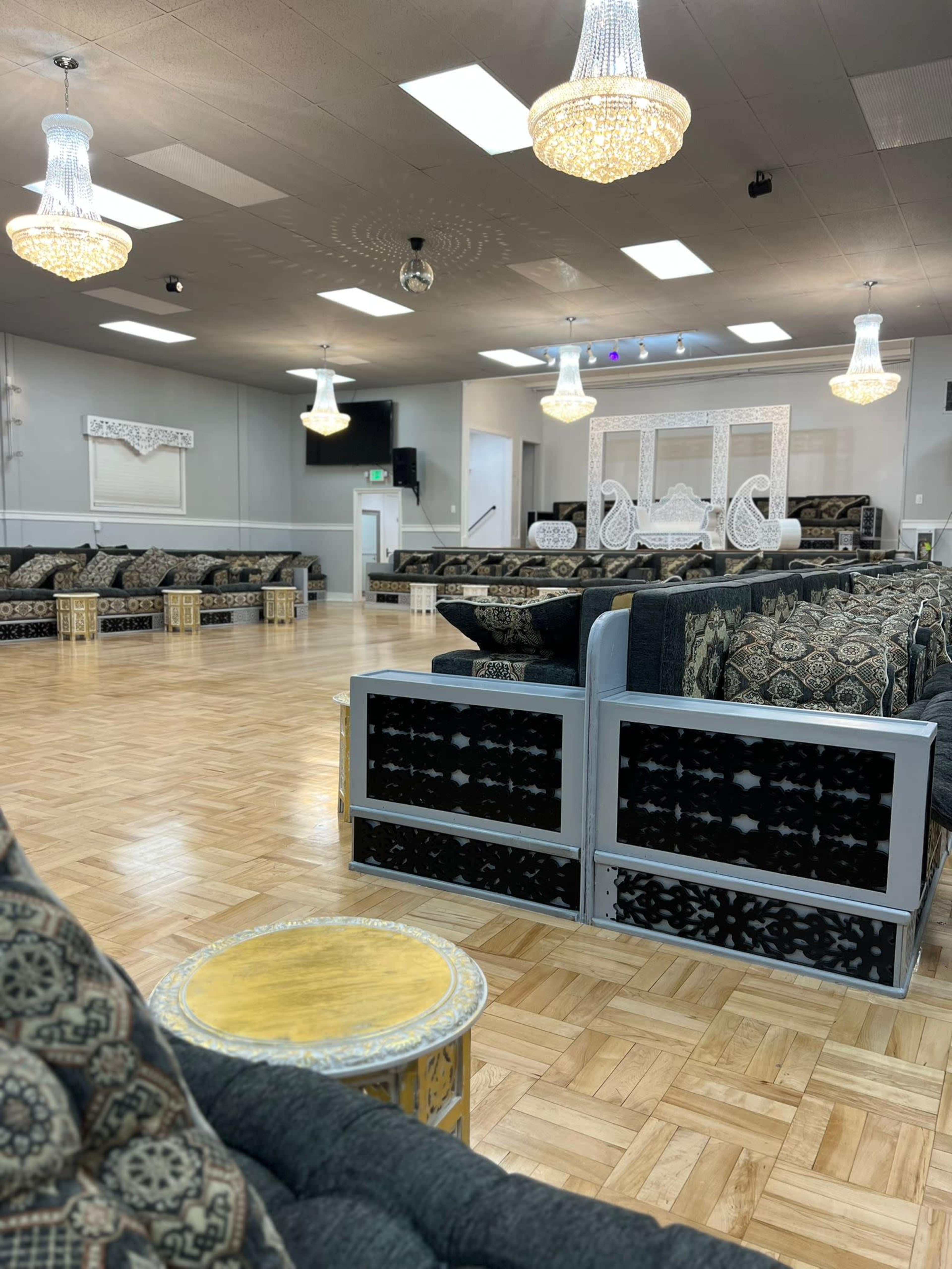 A spacious event hall with ornate seating arrangements, decorative chandeliers, and a polished wooden floor.