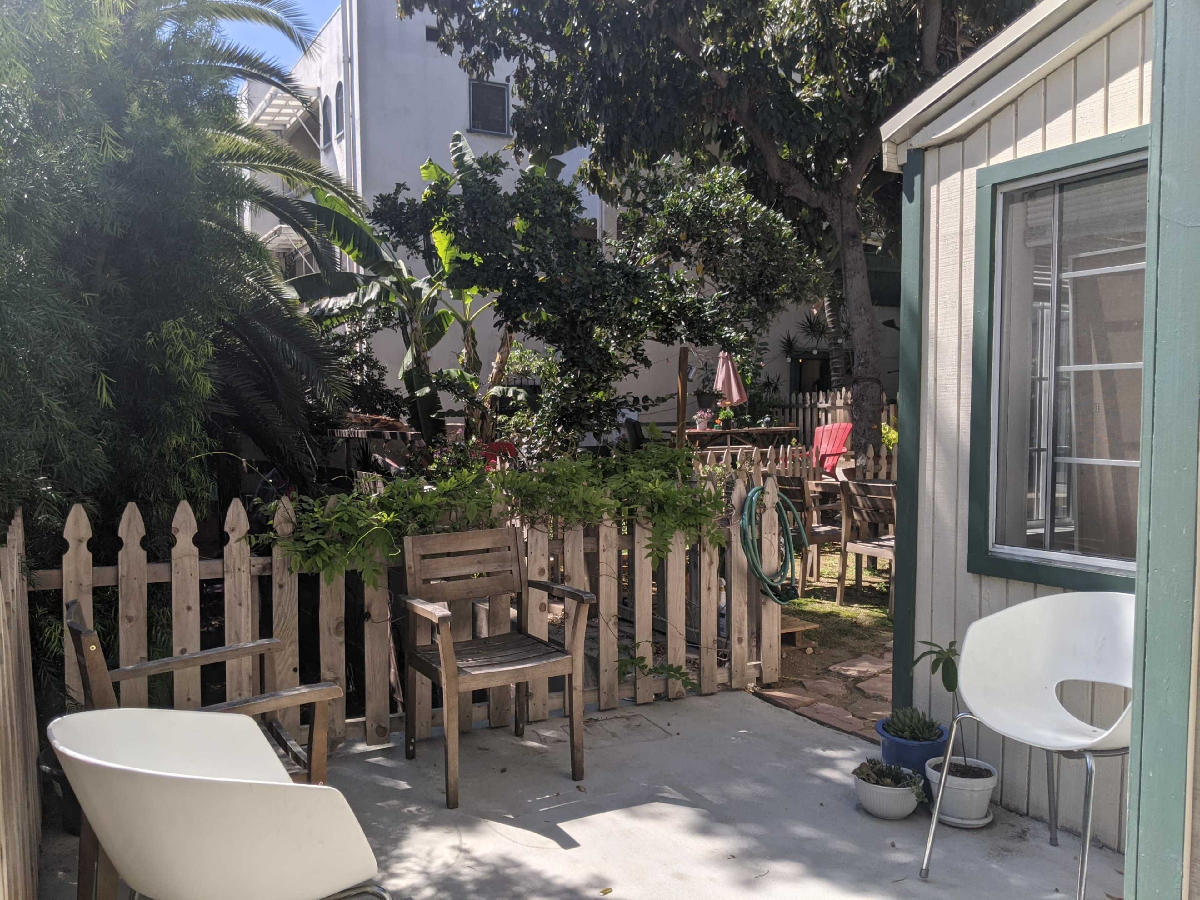 The image shows a small outdoor patio area with wooden furniture, a picket fence, and lush greenery surrounding the space.