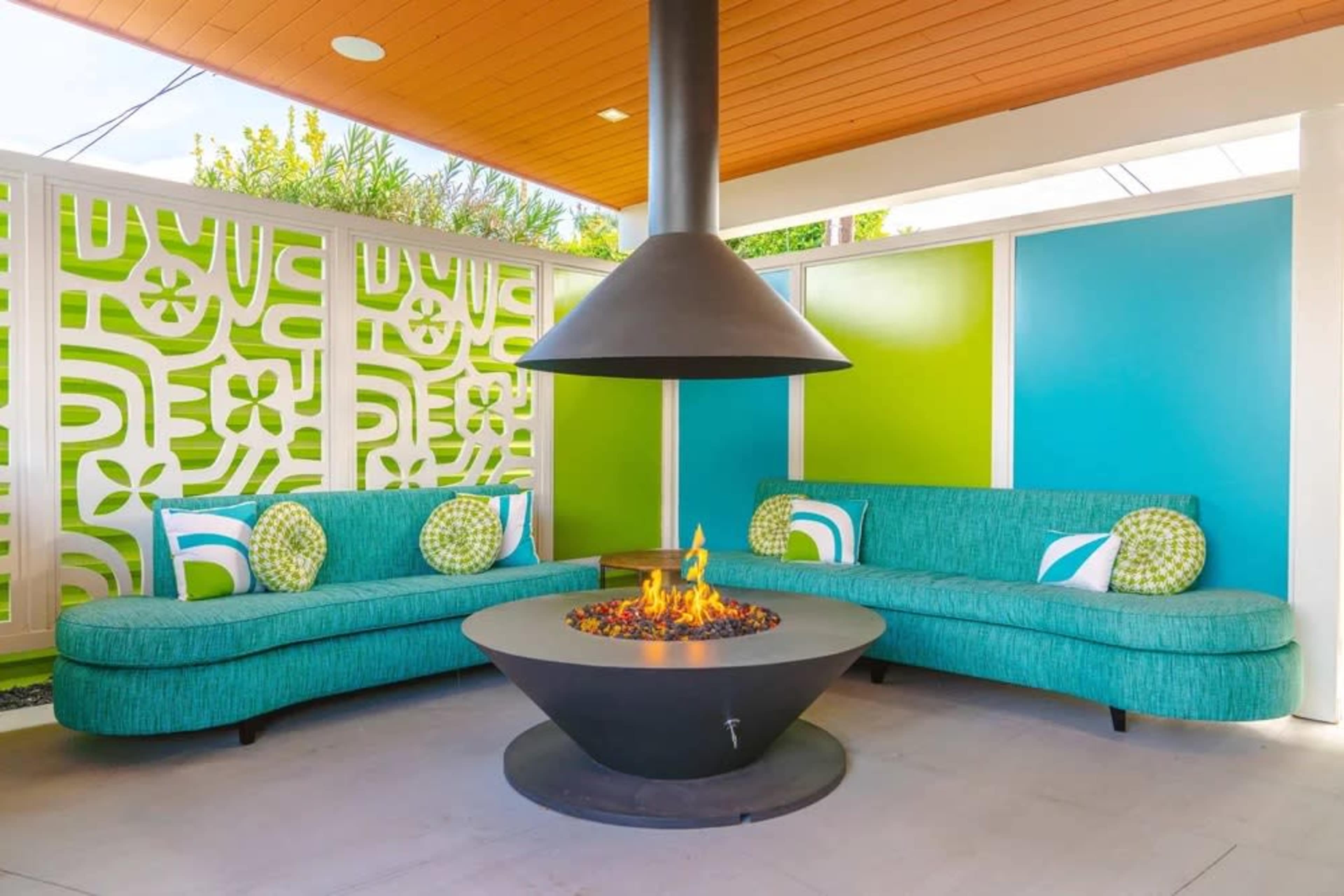 The image shows a cozy outdoor seating area featuring two teal sofa sections around a modern fire pit, with vibrant green and white patterned walls in the background.