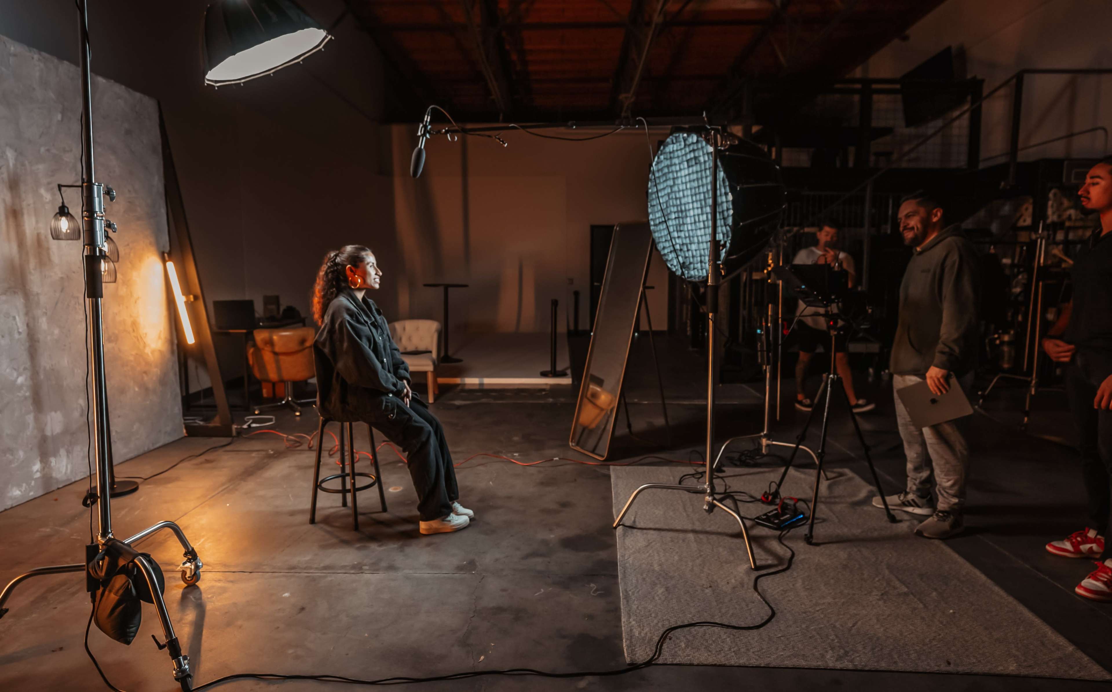 A person sits on a stool in a photography studio, surrounded by lighting equipment and crew members preparing for a shoot.