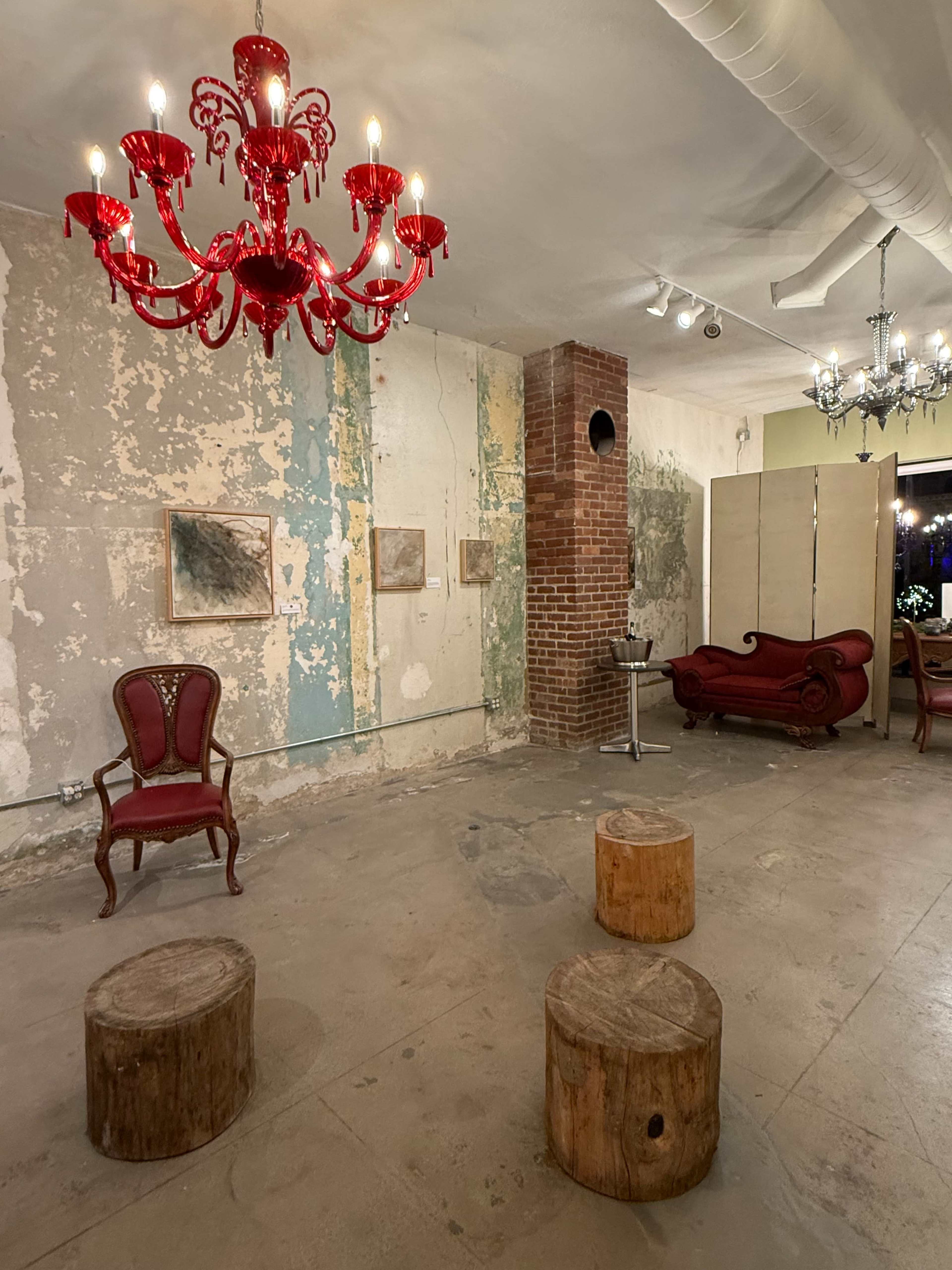 The image shows an art gallery with a red chandelier, vintage seating, and rustic wooden stools on a bare concrete floor against a backdrop of peeling walls.