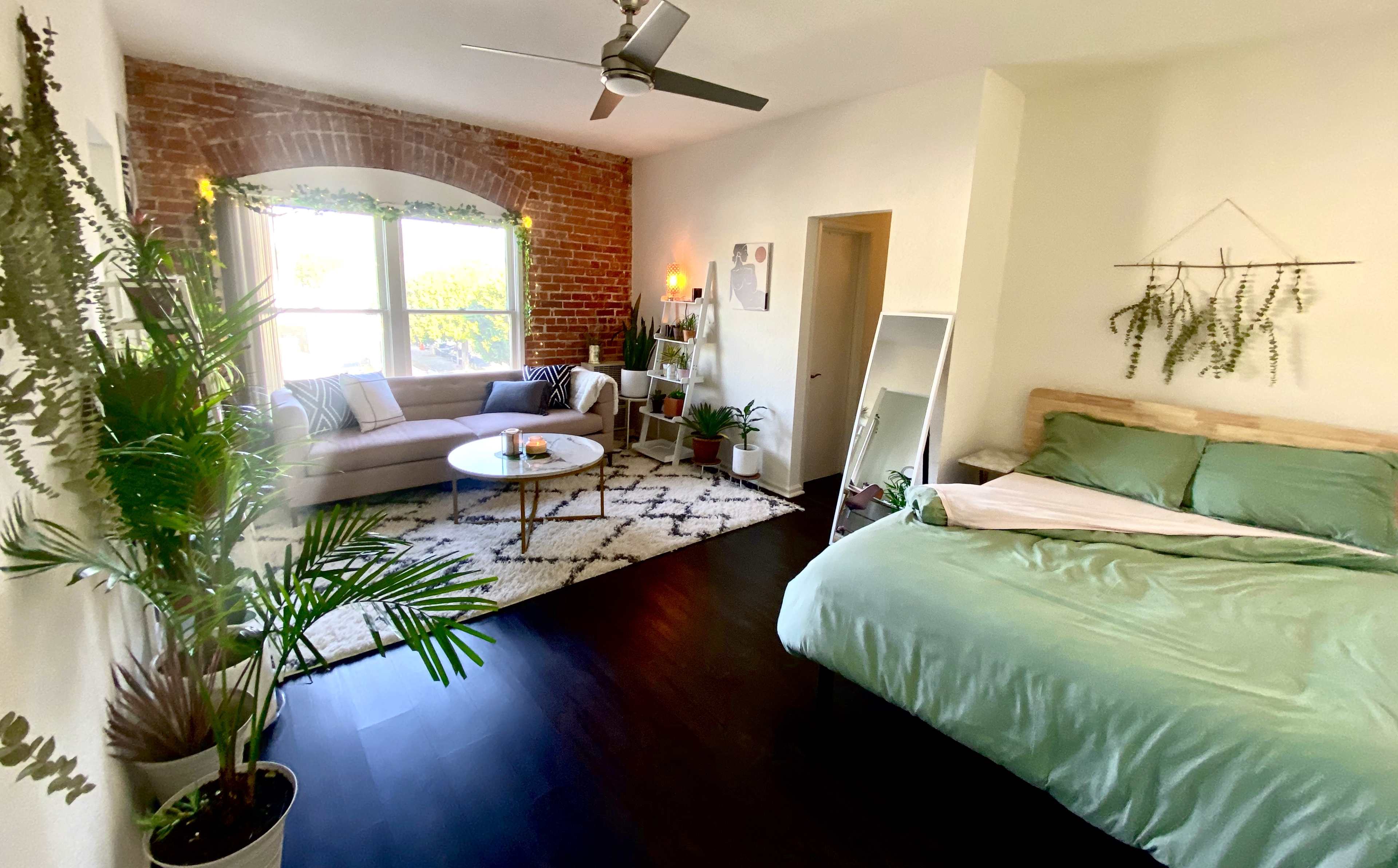 A cozy bedroom with a green bedspread, a sofa, a round table, and indoor plants surrounding a window with exposed brick walls.
