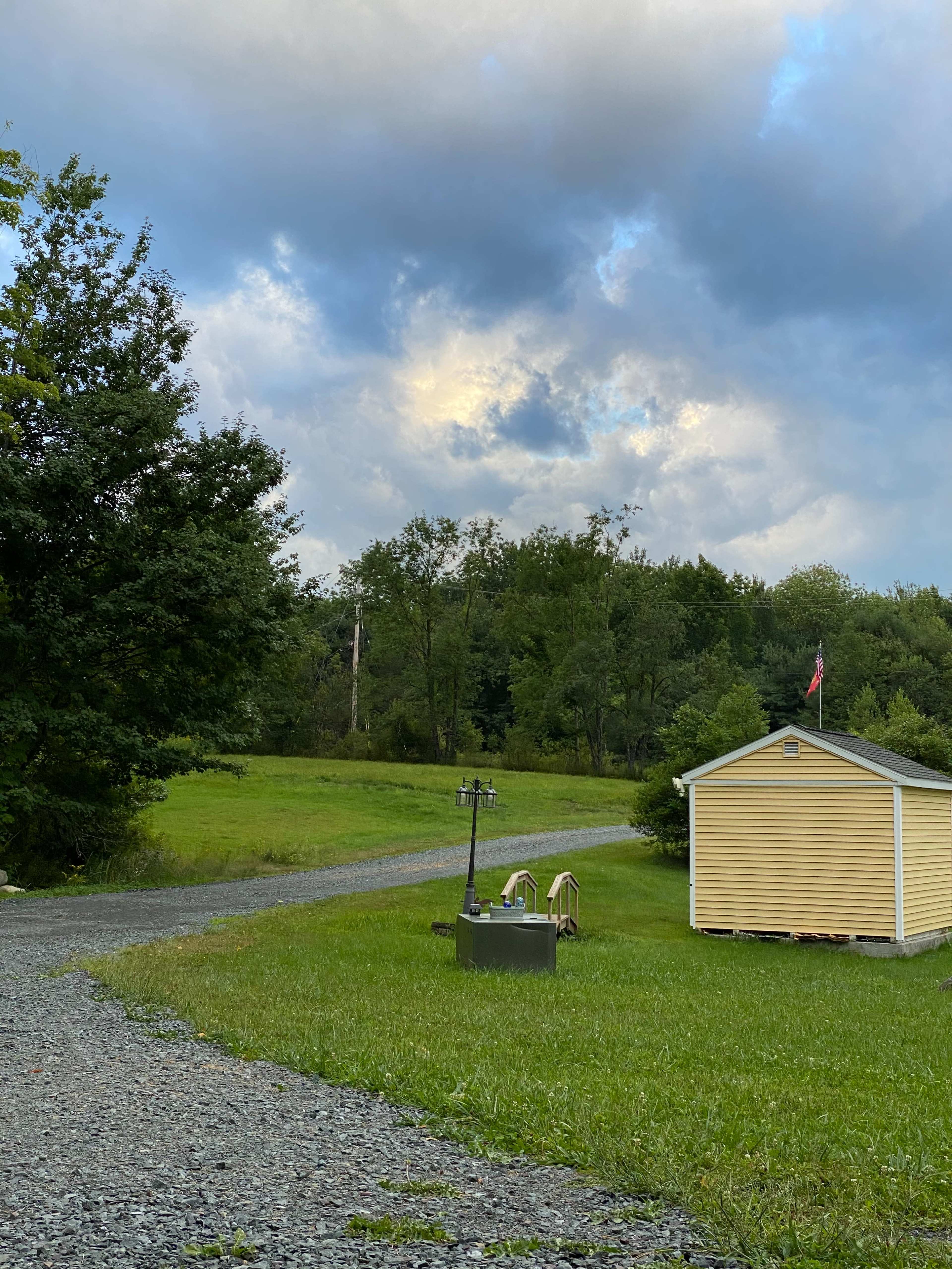 A gravel path leads to a small yellow shed surrounded by green grass and trees, under a cloudy sky.