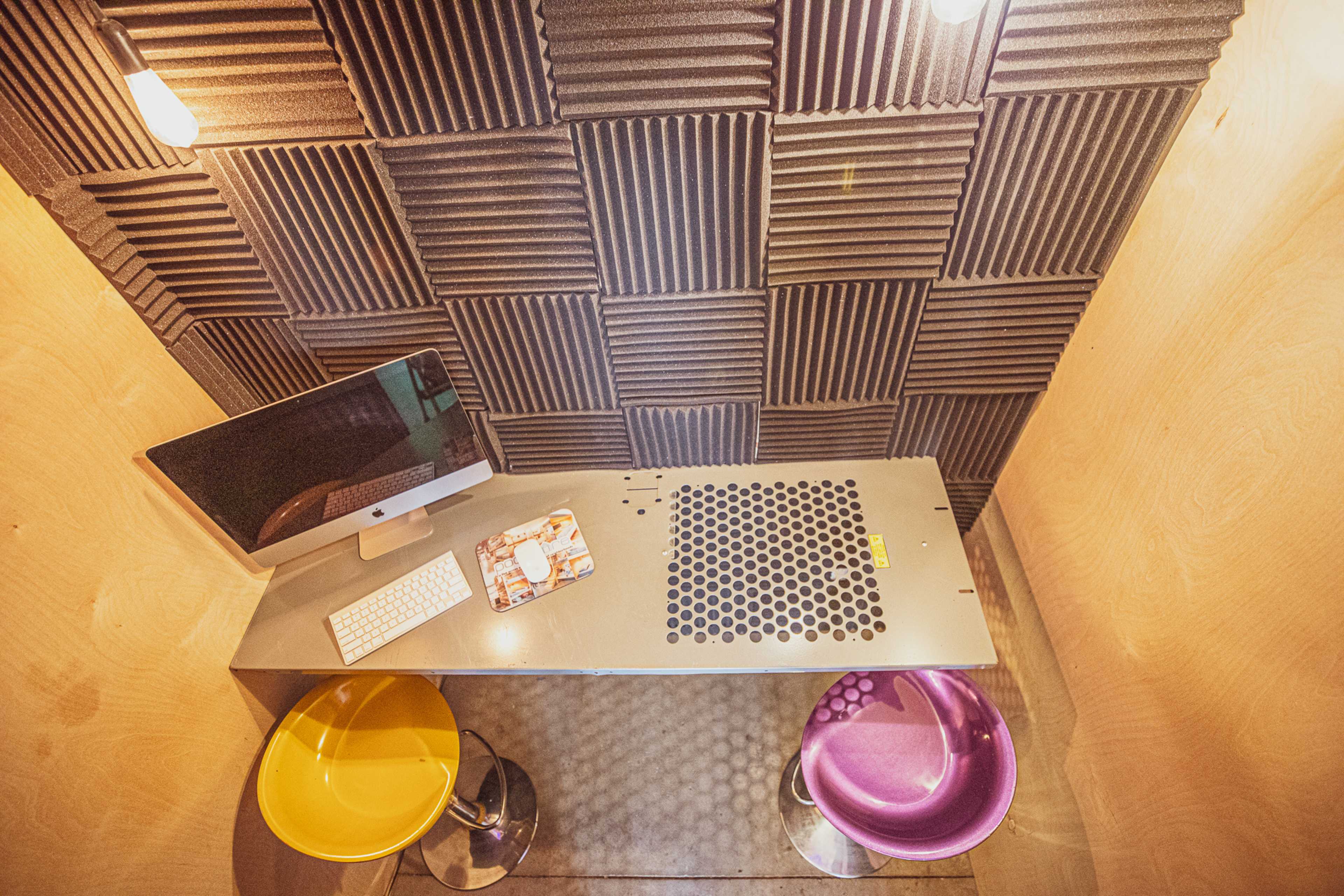 The image shows a small workspace featuring a computer on a metal desk with two colorful chairs and soundproofed walls.