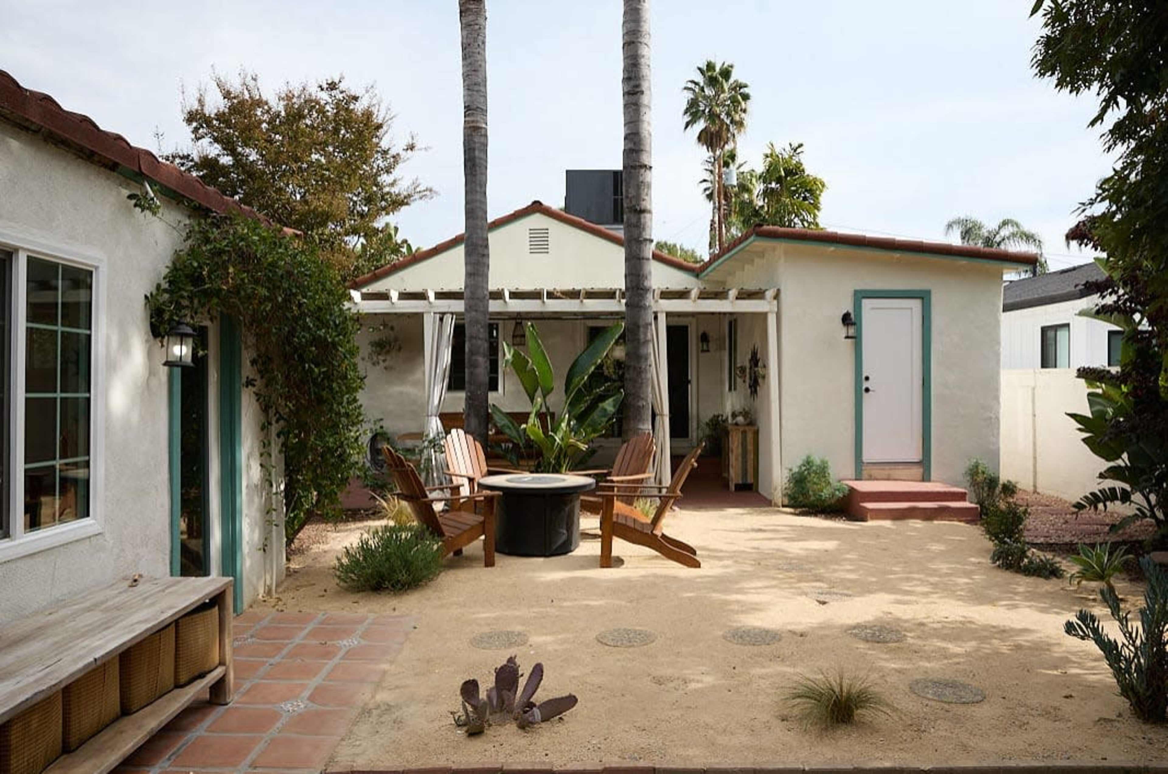The image shows a backyard with a sandy area, two palm trees, a seating arrangement with wooden chairs around a fire pit, and a house with light-colored walls and green accents.