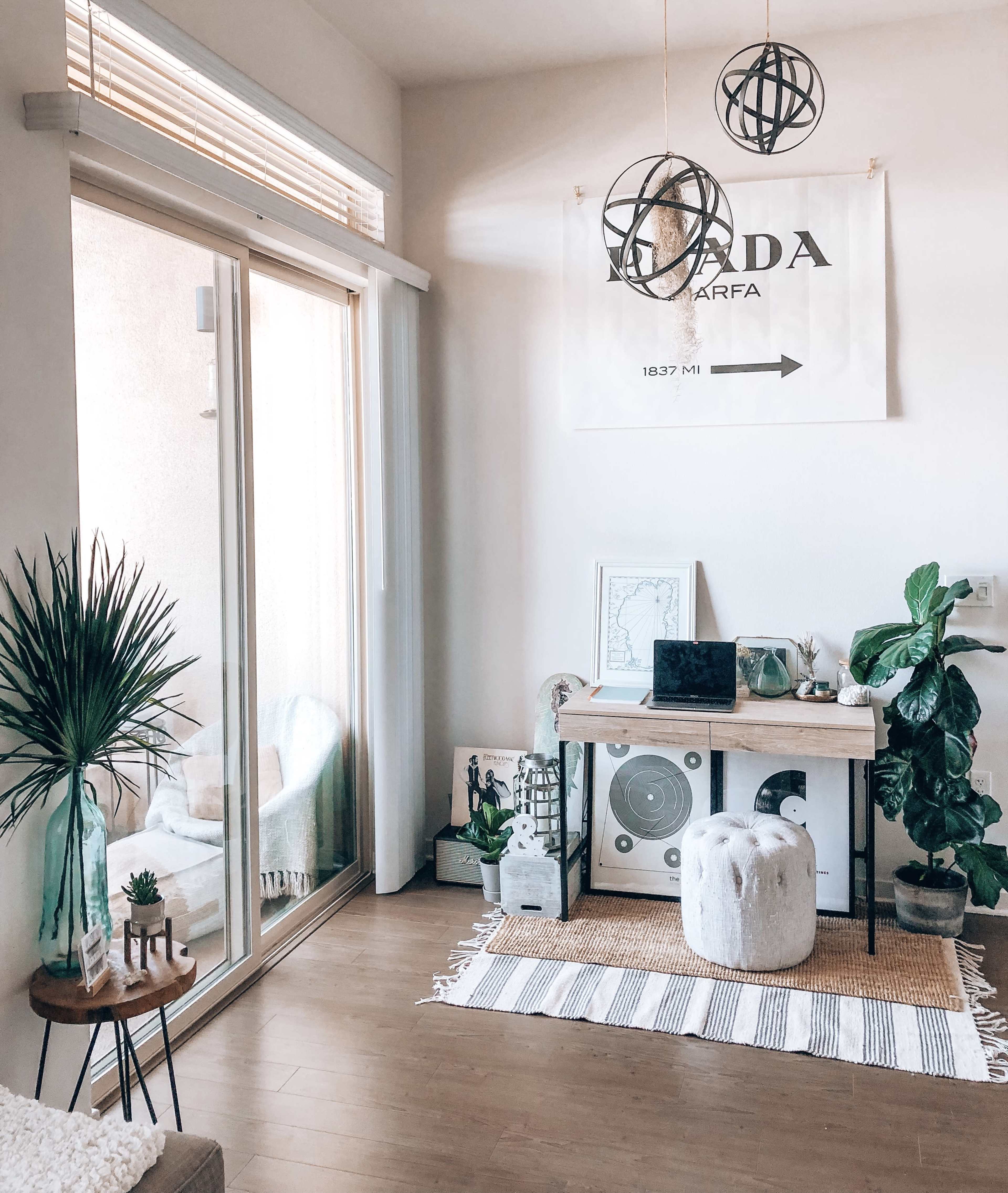 A bright and airy room featuring a workspace with a laptop on a table, surrounded by plants and decorative elements, along with a large window that allows natural light to enter.