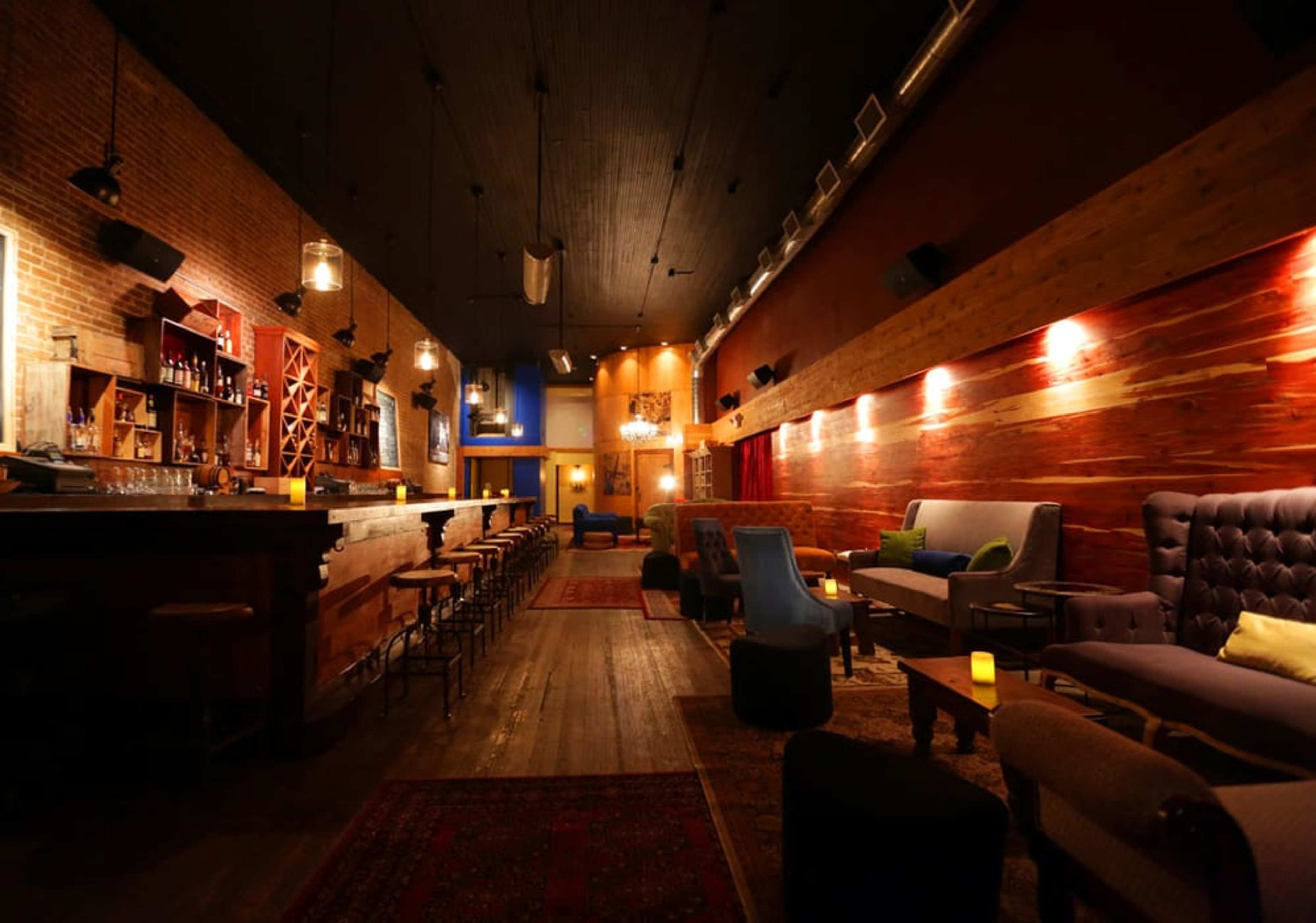 A dimly lit bar with a wooden interior, featuring a long bar counter, seating areas with plush chairs, and decorative lighting.