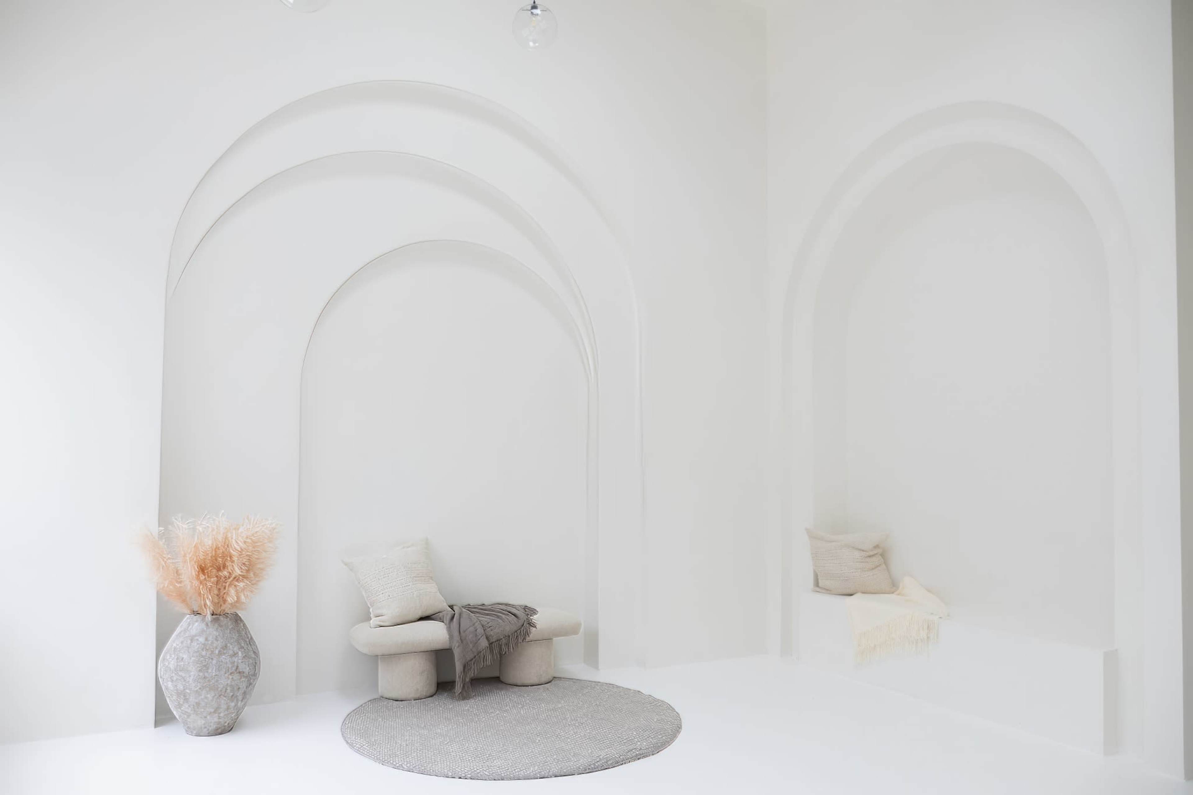 A minimalist interior with archways, a soft rug, a sculptural seating arrangement, and a vase with dried foliage.