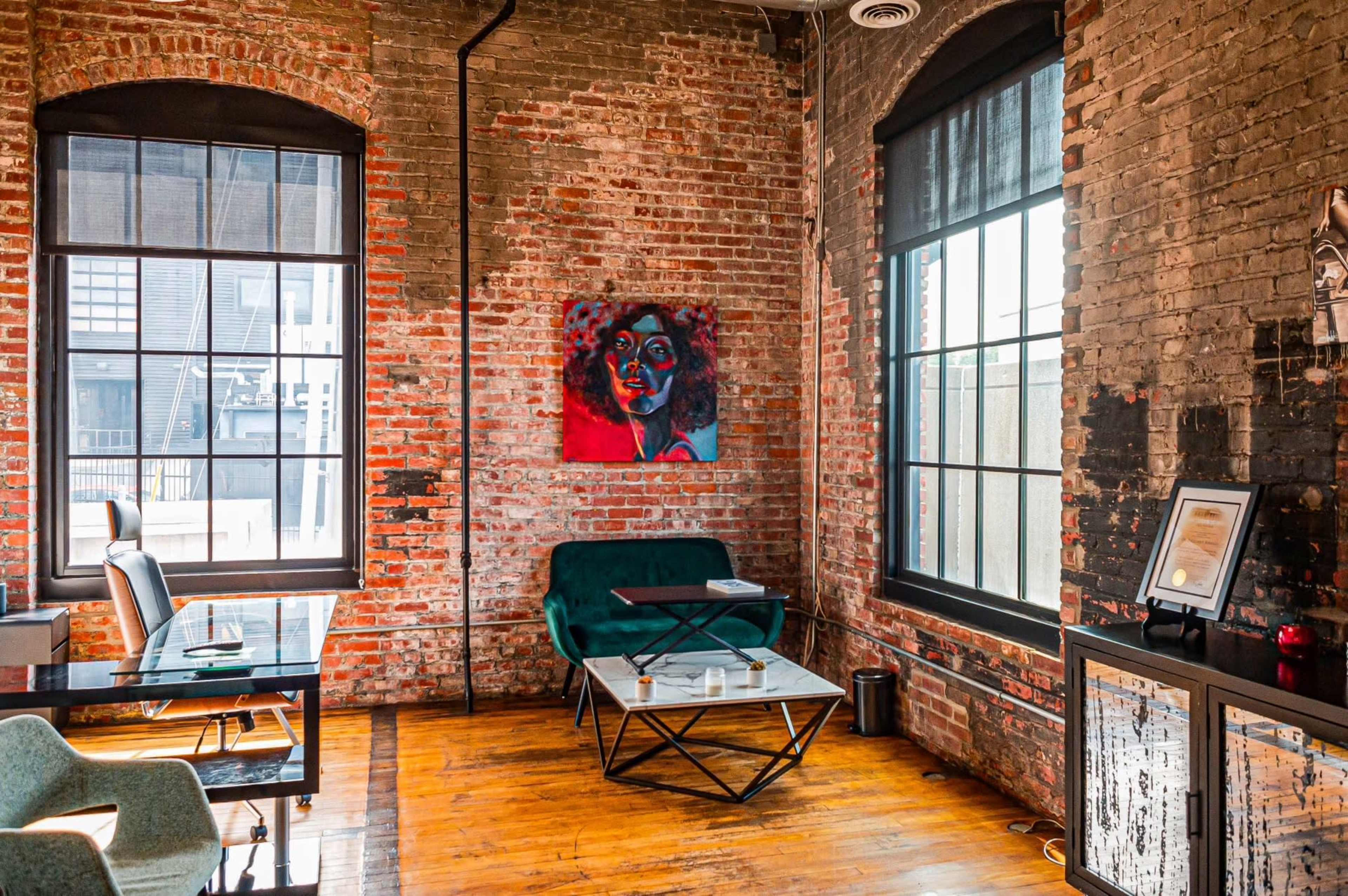 A corner of a room features exposed brick walls, large windows, a green sofa, a glass coffee table, and a desk with a chair.