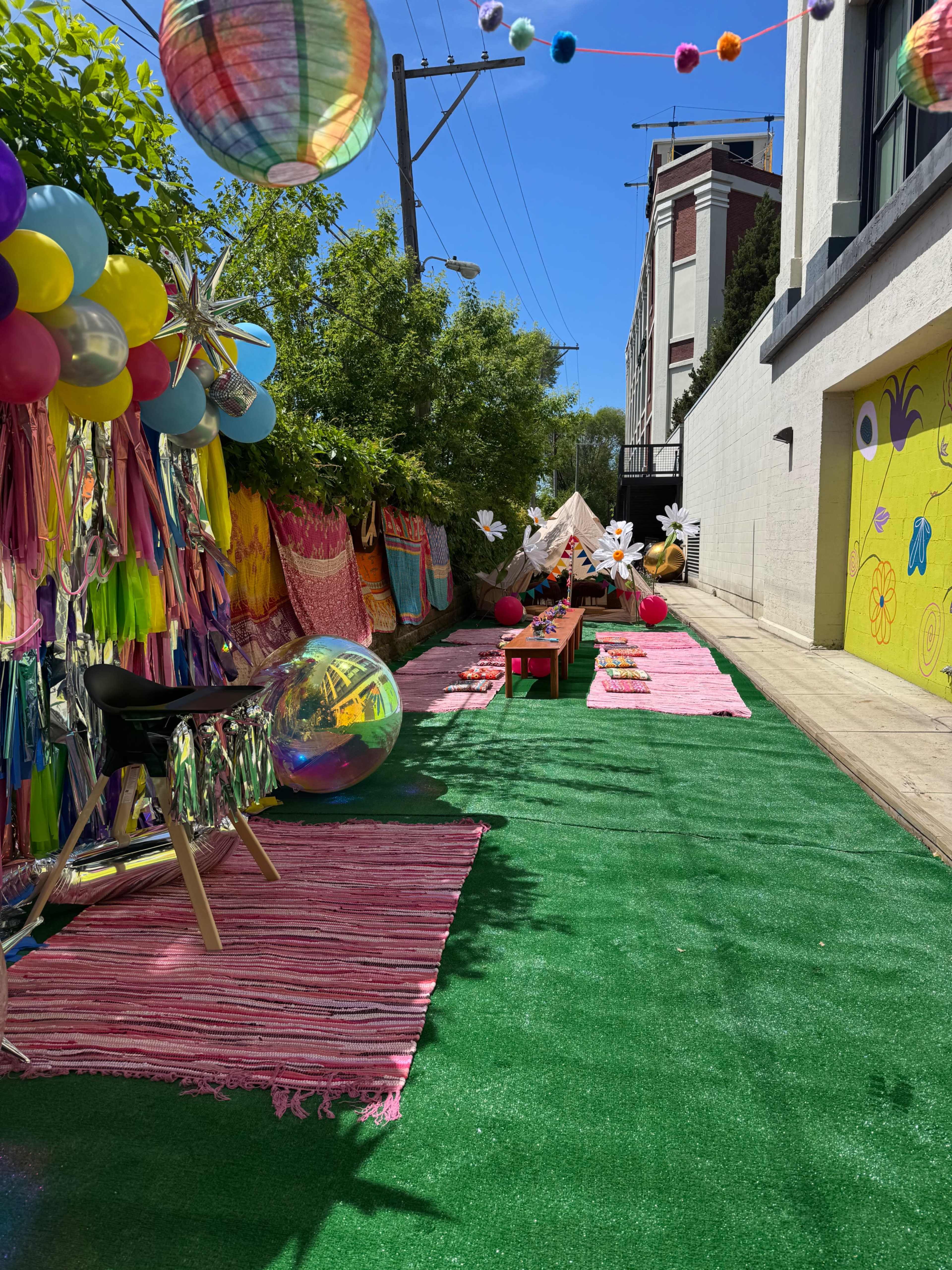 The image shows a colorful outdoor alleyway decorated with balloons, streamers, and a green carpet, featuring tables set for a gathering.