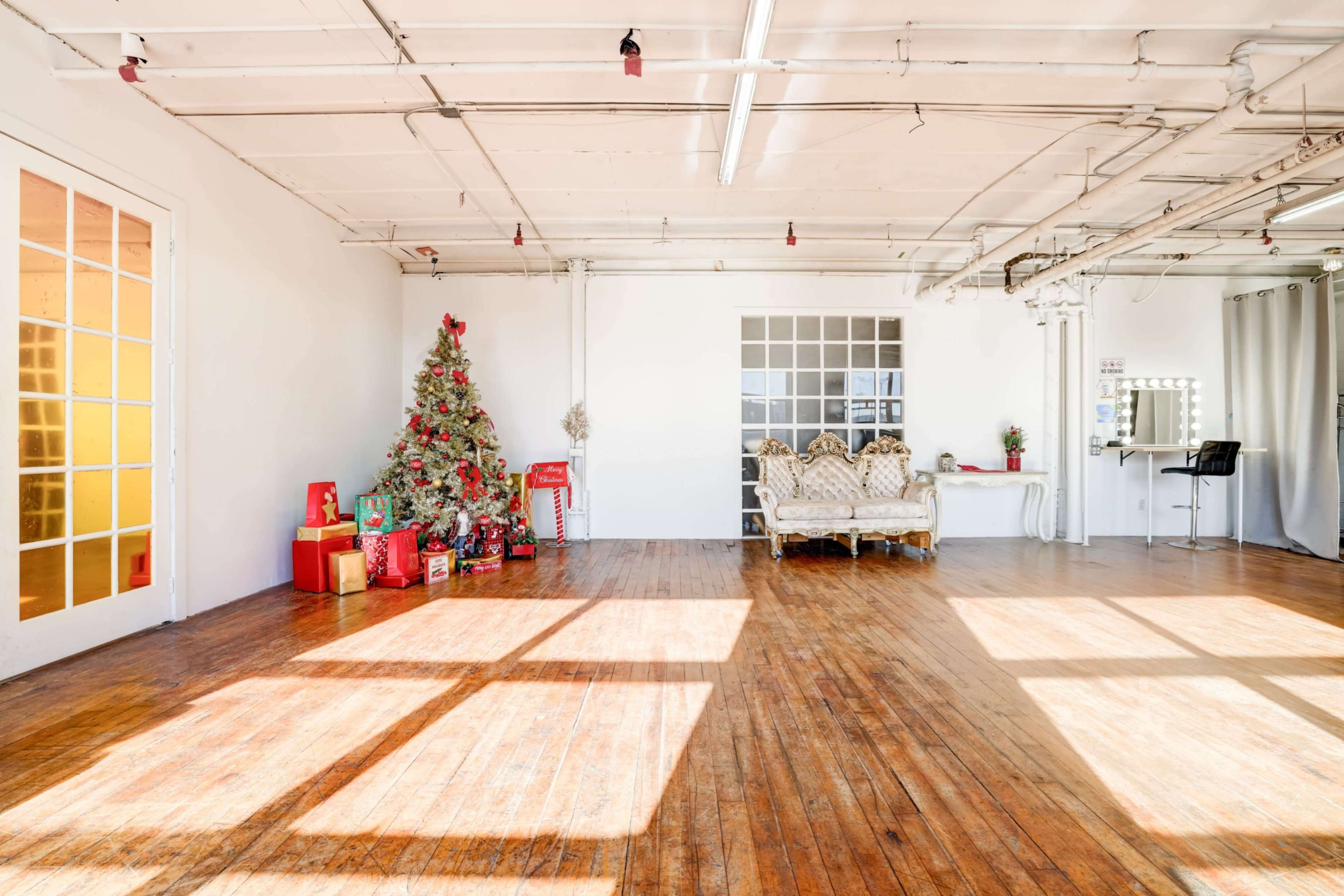 A spacious, well-lit room features a Christmas tree adorned with decorations, a sofa, gifts, and a mirror setup.