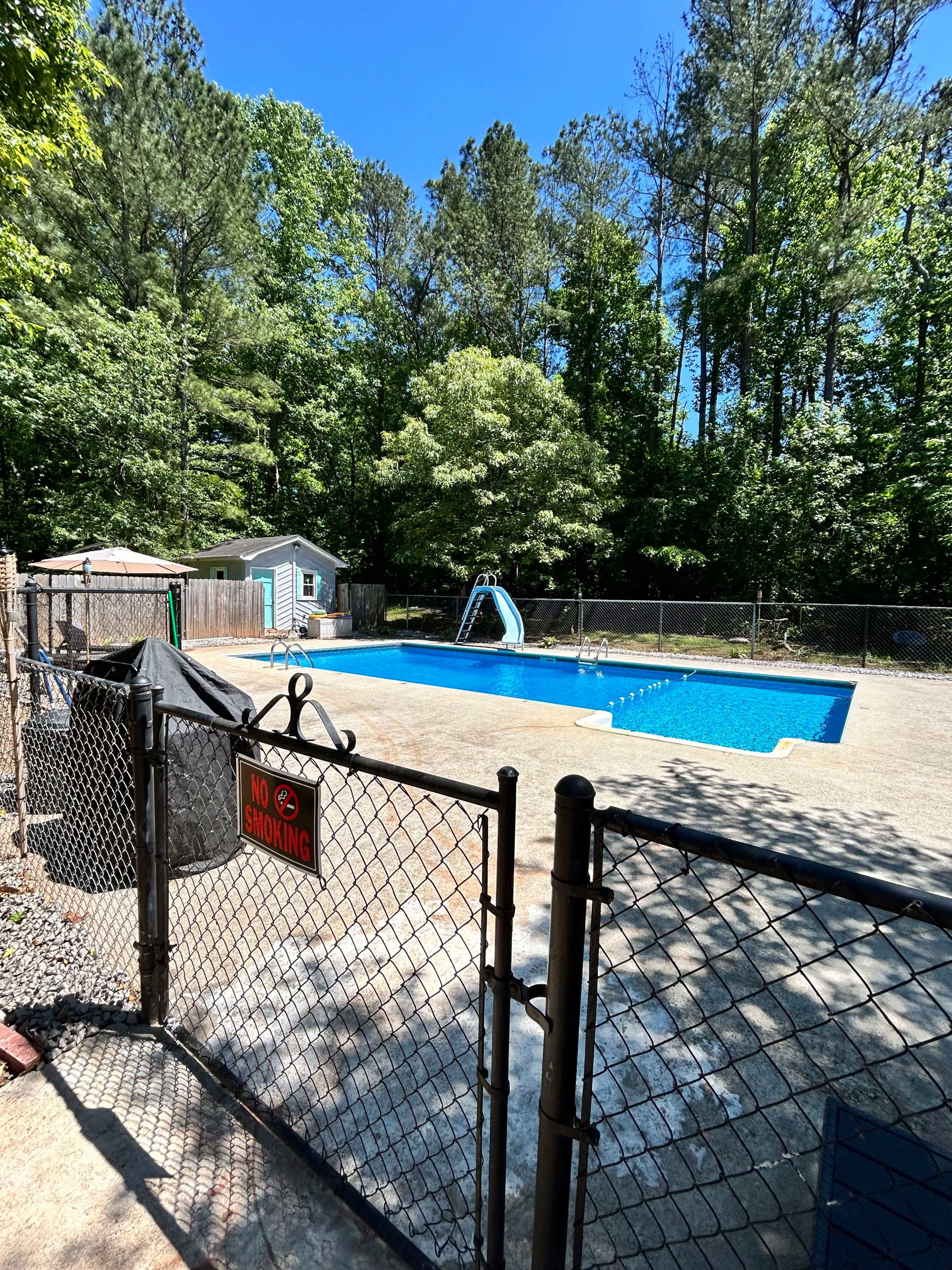 A fenced swimming pool area with a blue pool, a slide, and surrounded by trees.