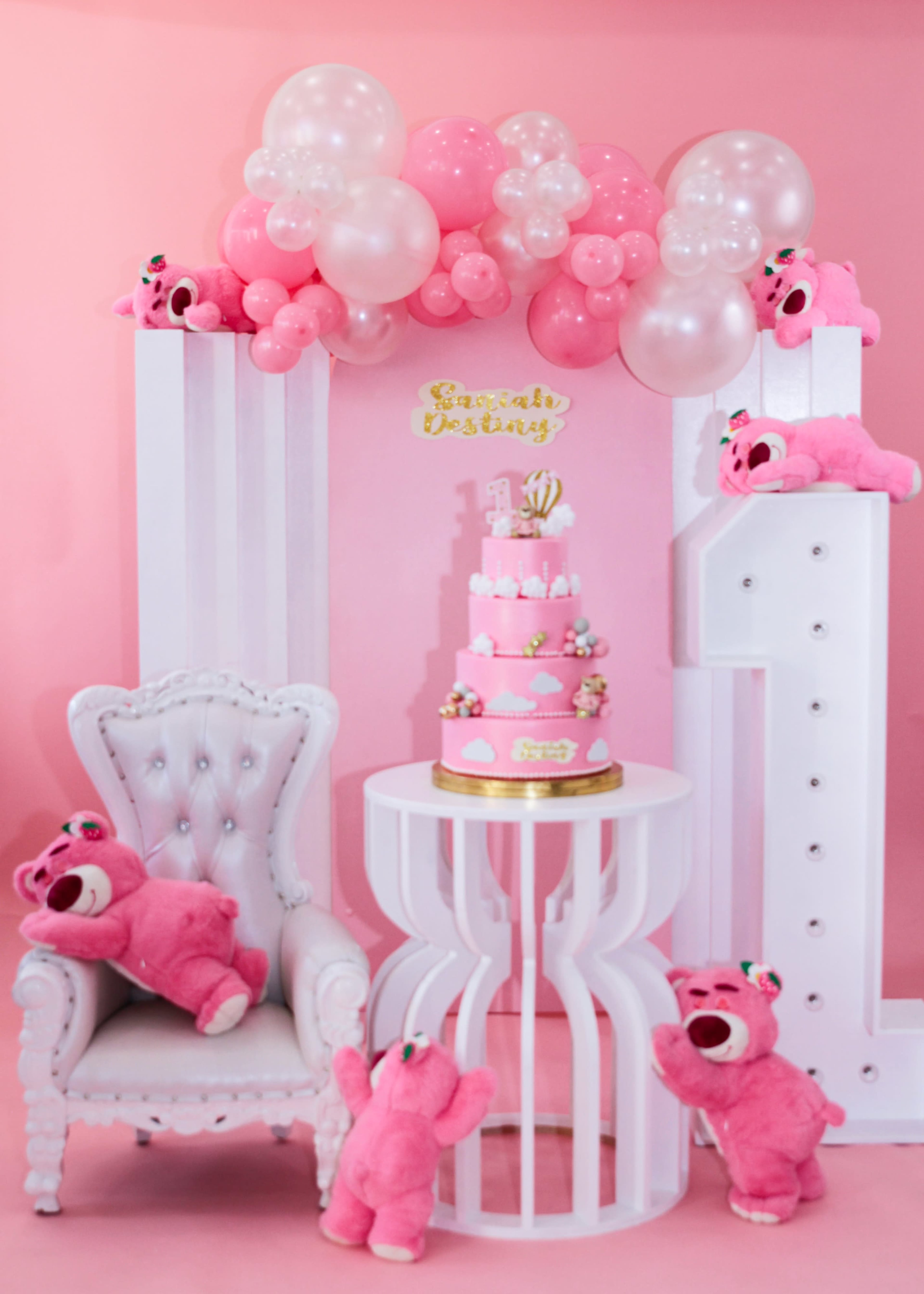 A pink-themed birthday setup features a large cake on a table surrounded by plush toys and balloon decorations.