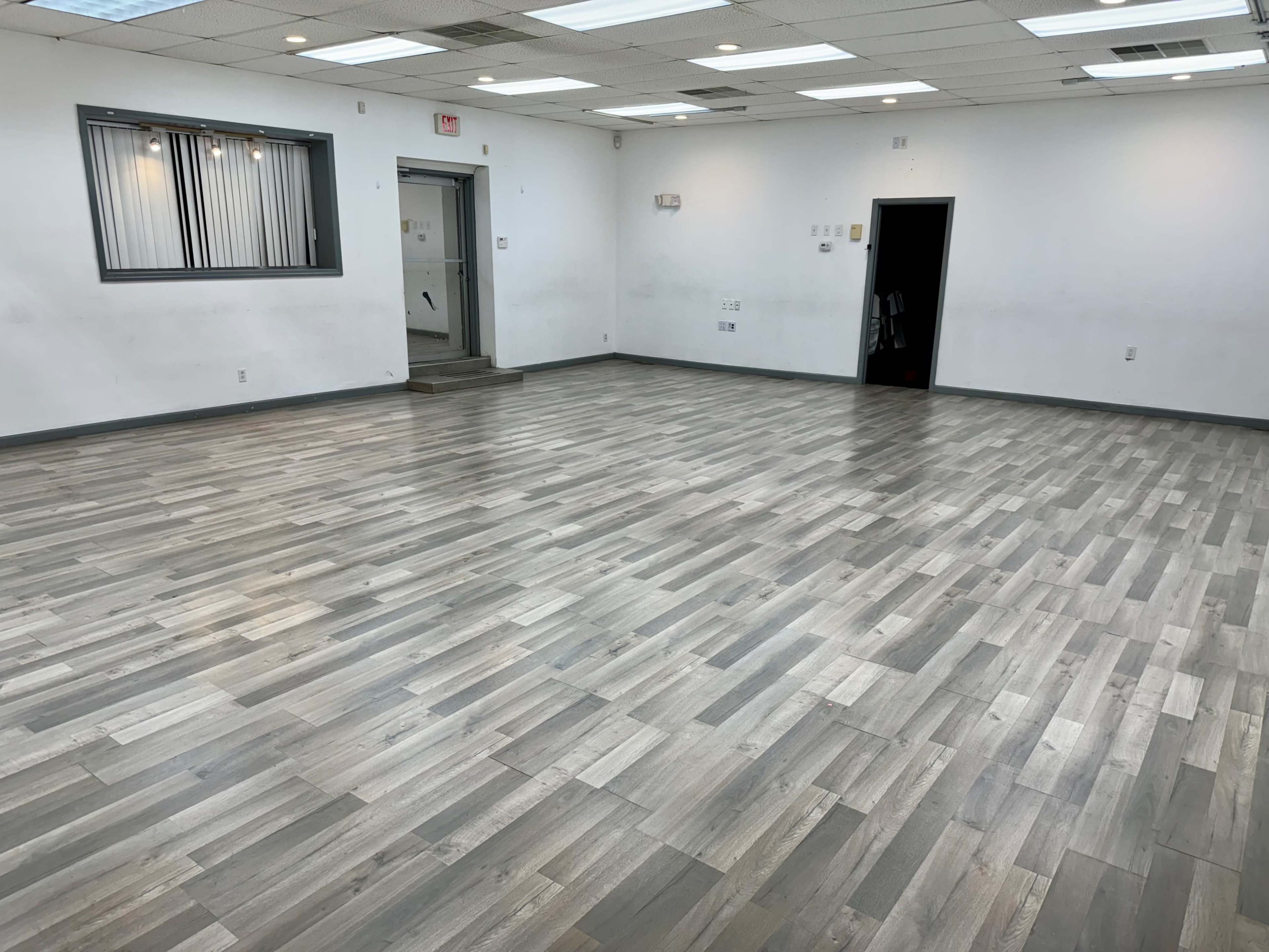 A large, empty room with laminate flooring and white walls, featuring two doorways and overhead lighting.