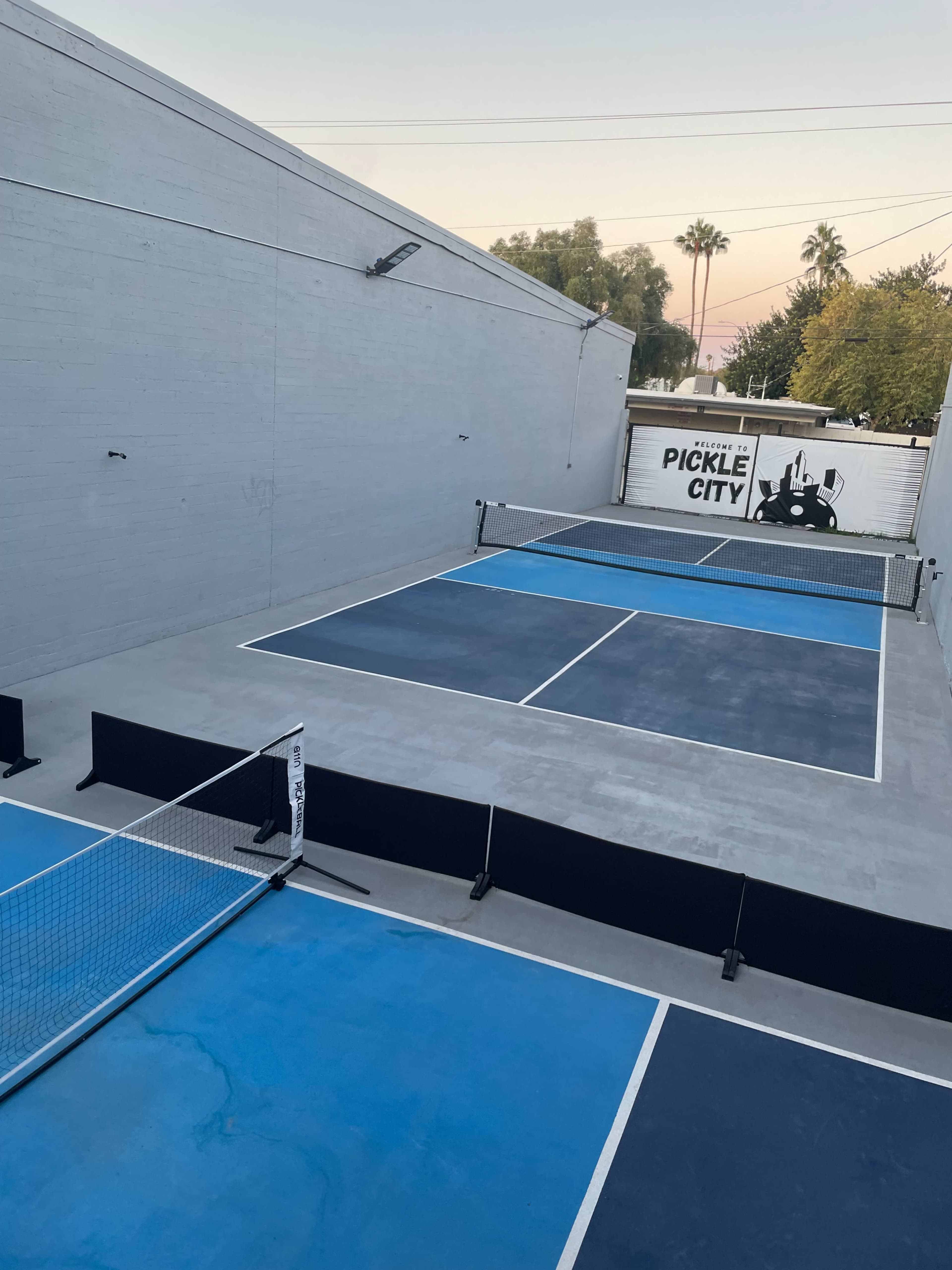 The image shows a pickleball court with blue and gray surfaces, surrounded by a fence, and a wall featuring the text "PICKLE CITY."