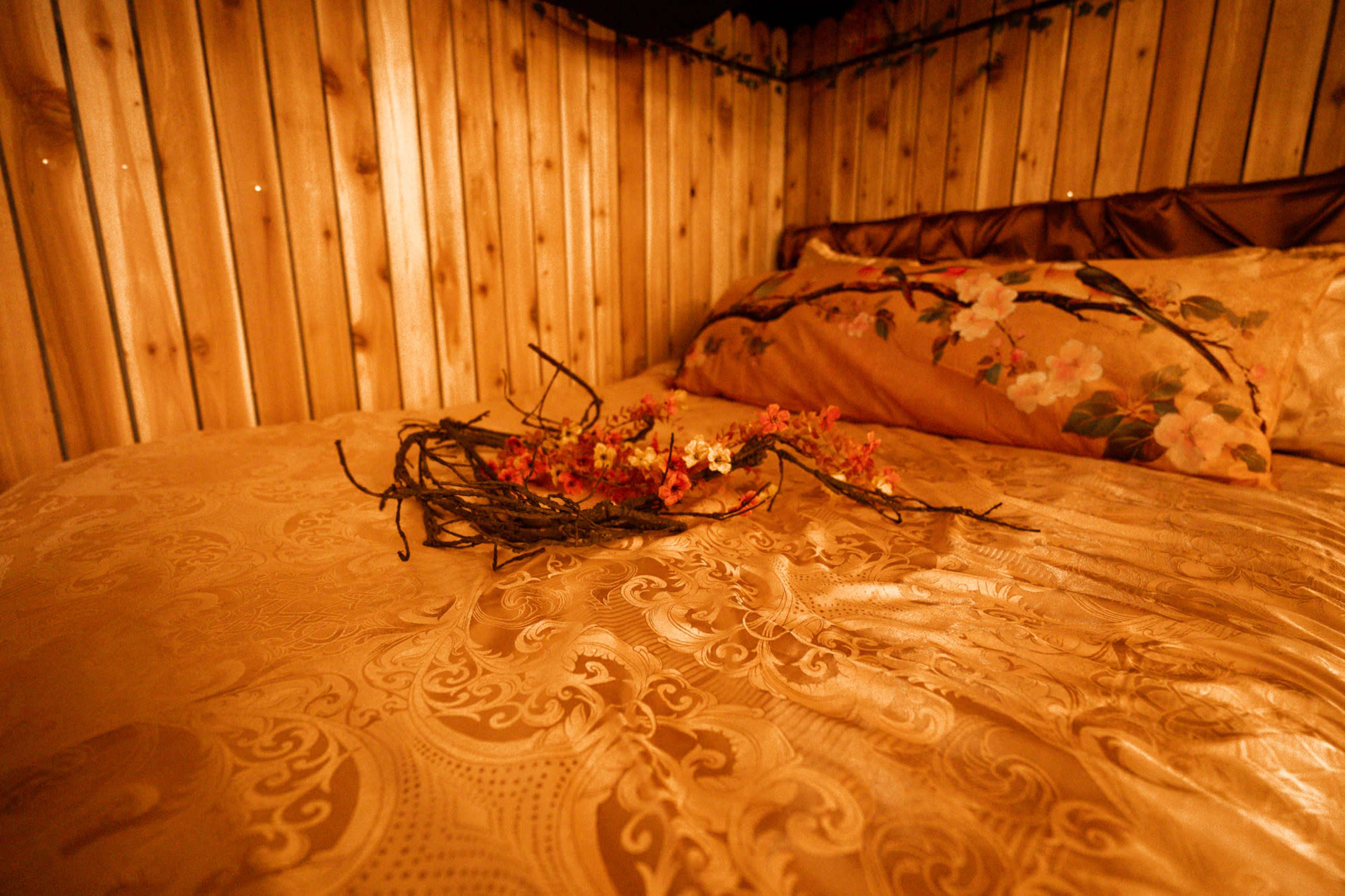 A bed with a decorative gold bedspread features a floral pillow and a wreath of dried flowers resting on top.