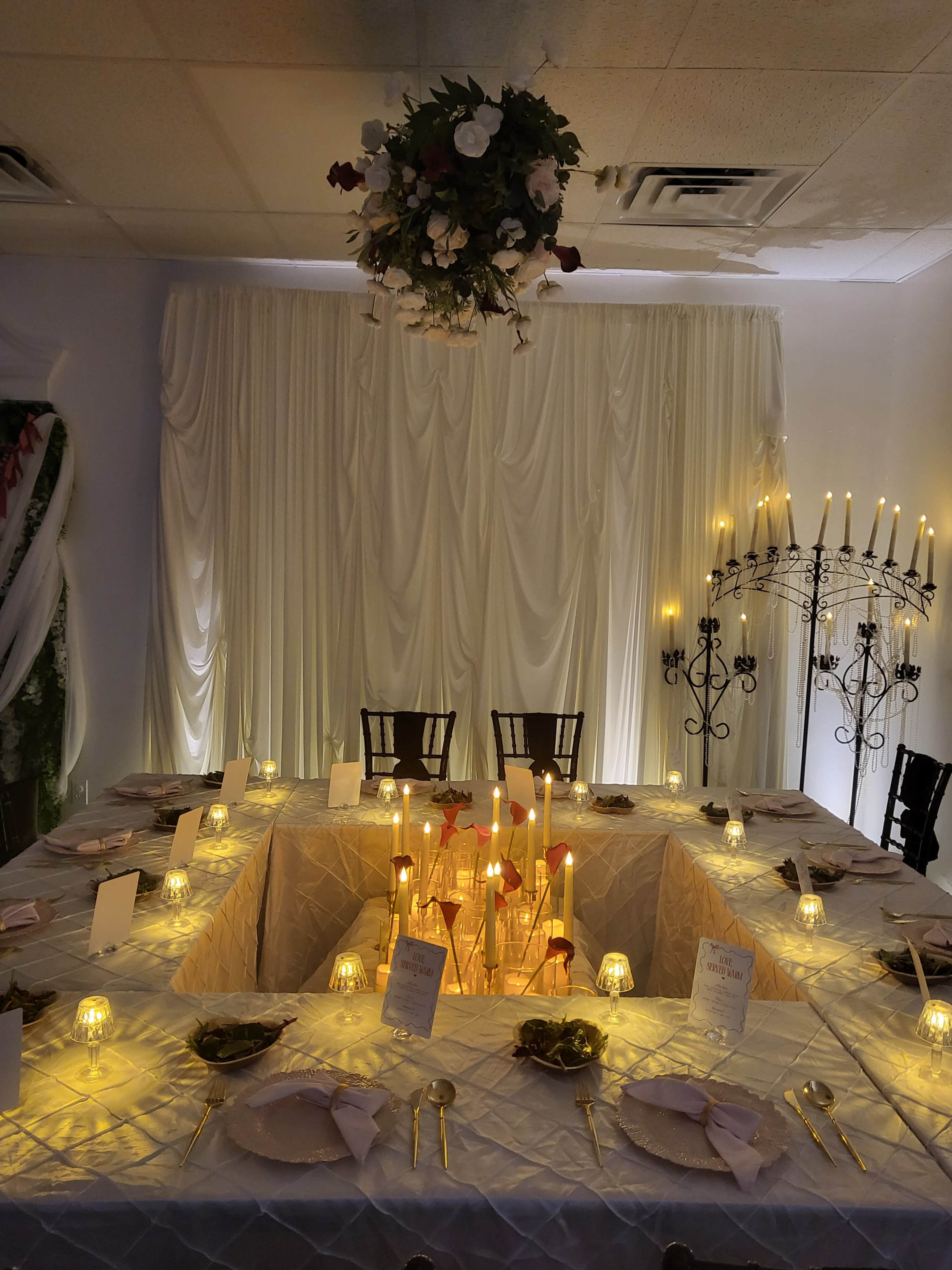 The image shows a decorated dining area with a large table set for a formal meal, surrounded by candles and soft lighting, beneath a floral arrangement hanging from the ceiling.