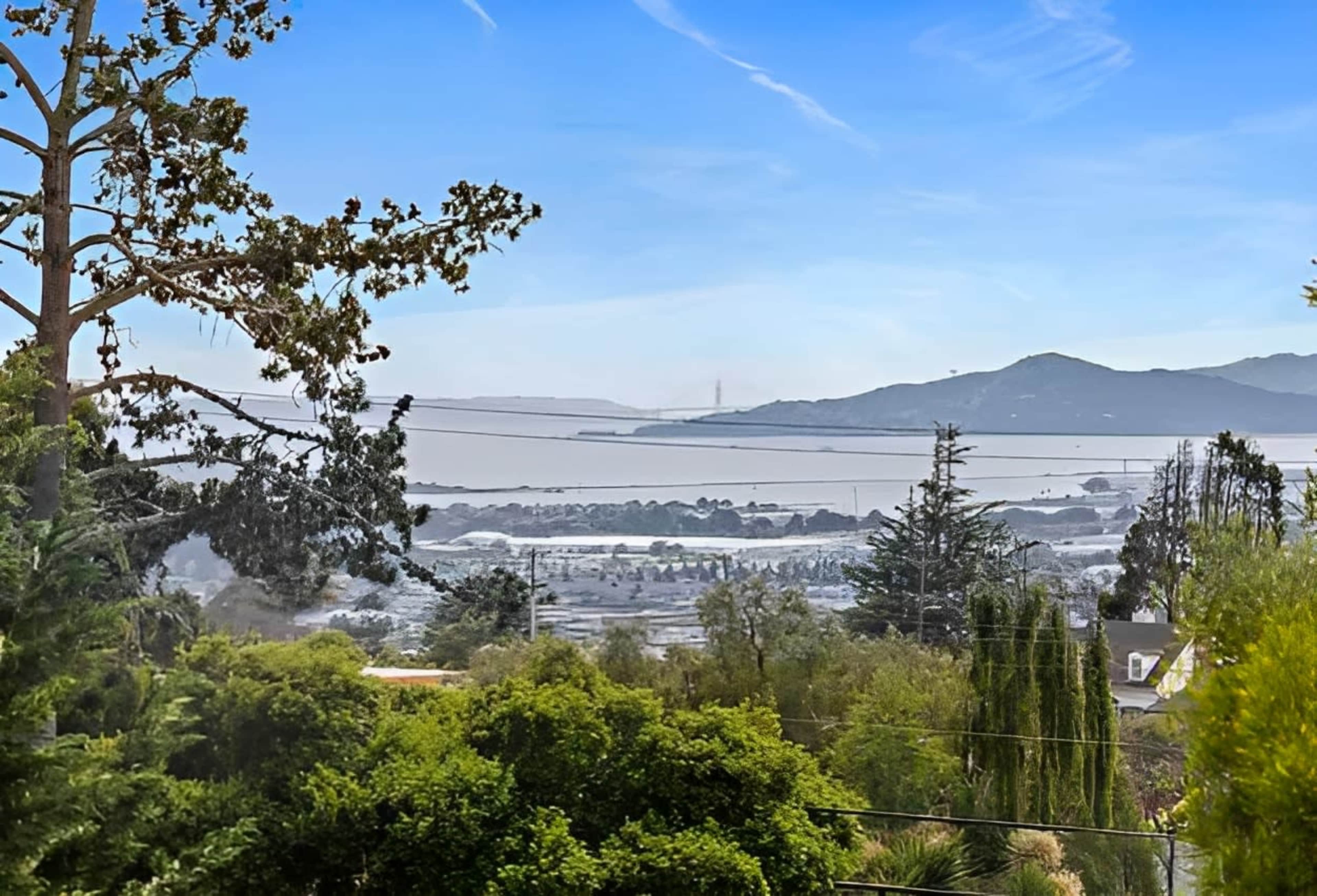 A view of a coastal landscape featuring trees, water, and distant hills under a clear blue sky.