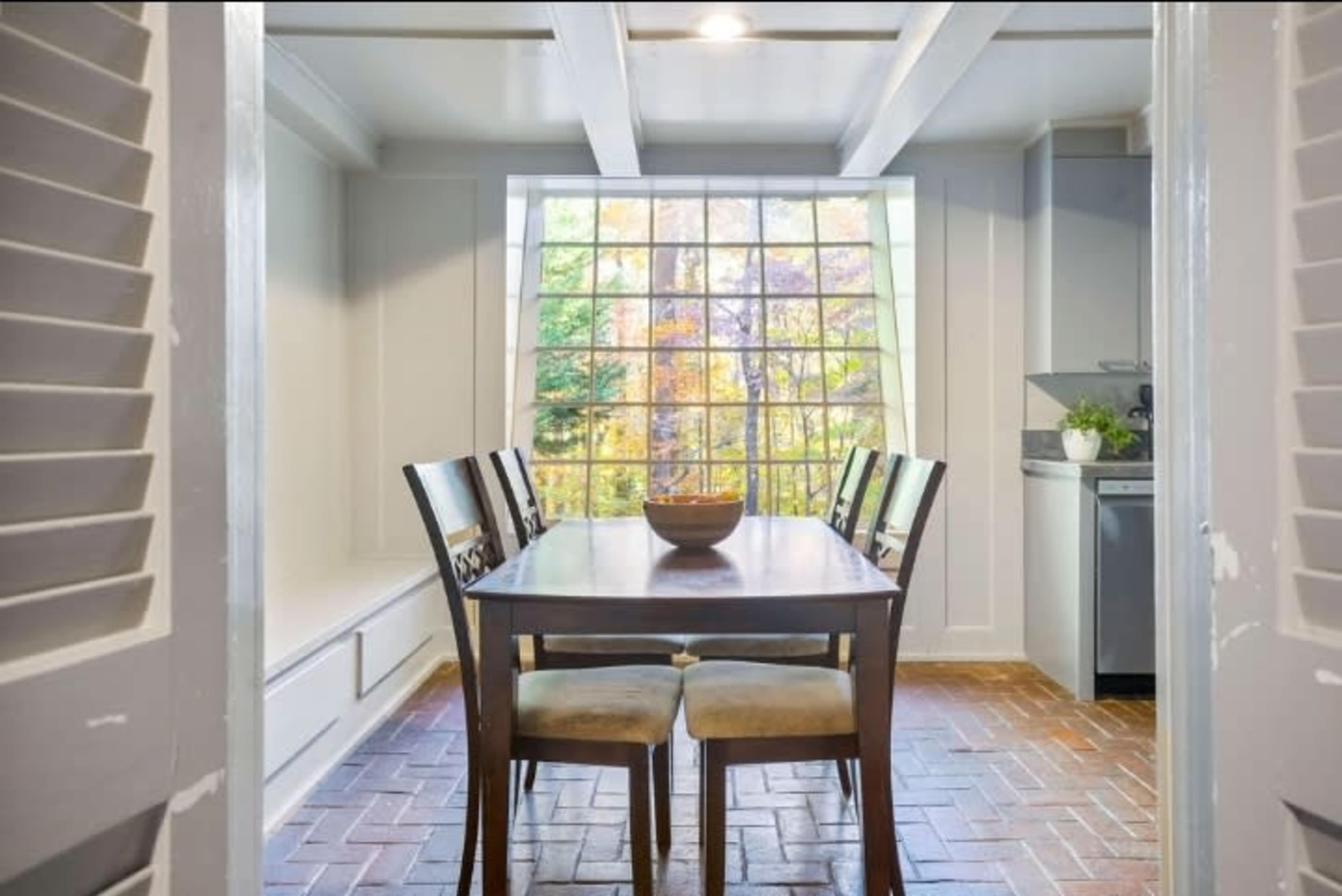 A dining area features a wooden table surrounded by four chairs, with a large window showing a view of colorful trees outside.