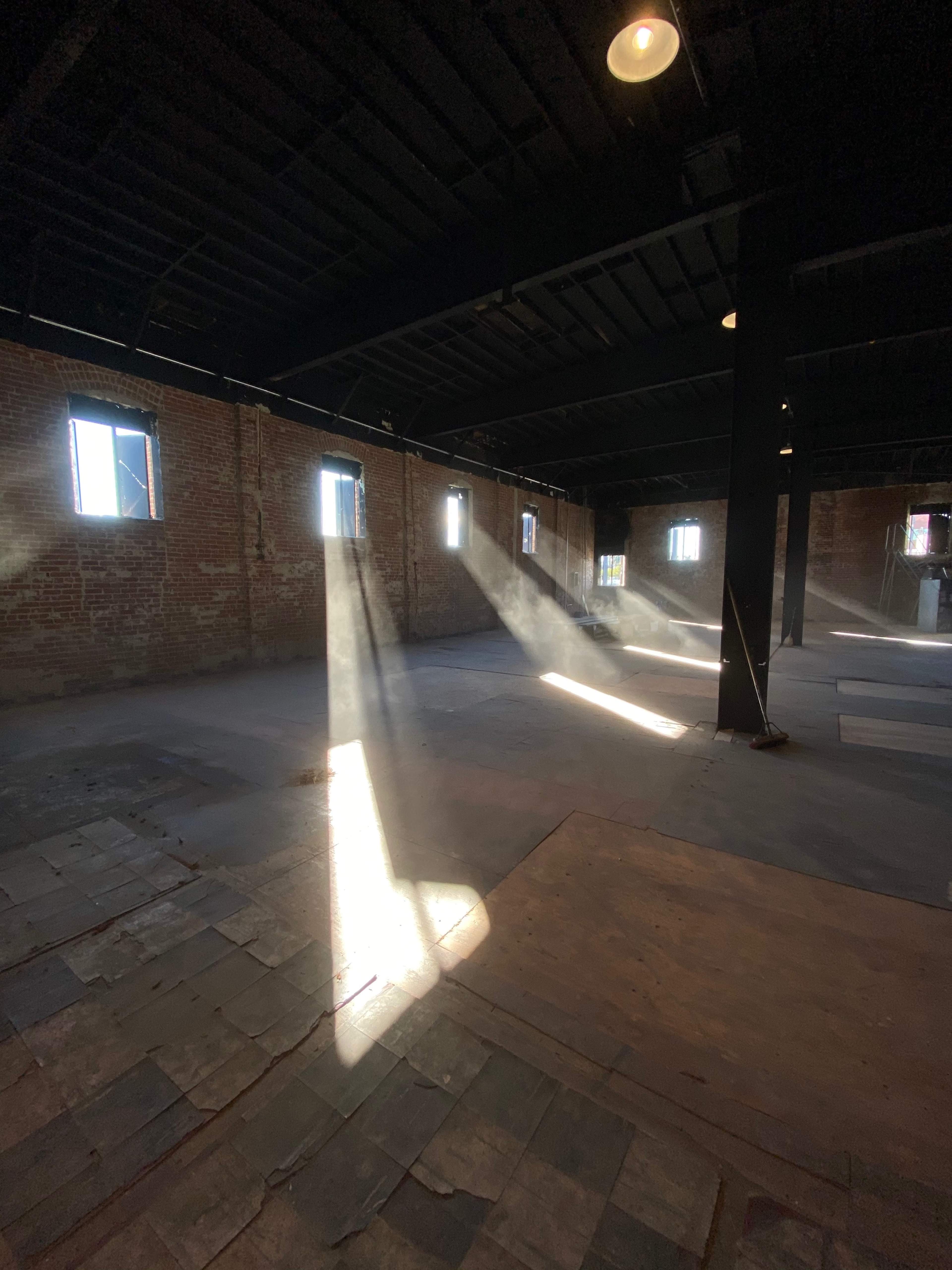 Sunlight streams through windows, illuminating dust particles in an empty brick-walled warehouse.