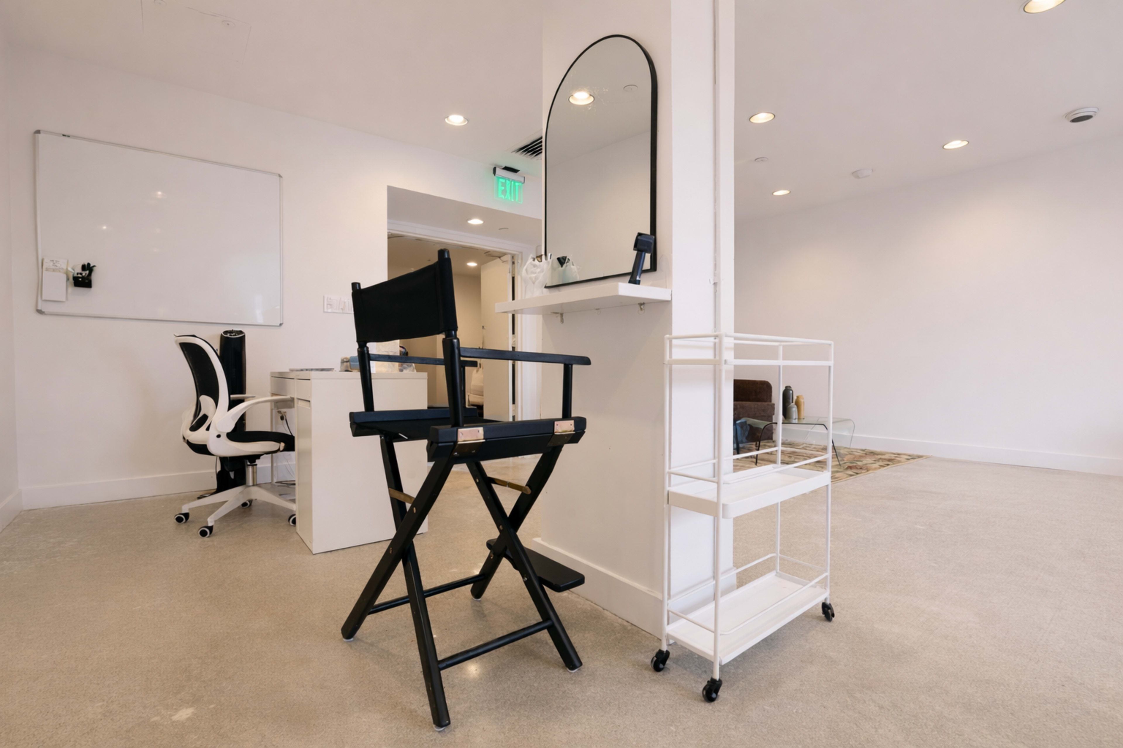 The image shows a minimalist room with a black director's chair, a circular mirror mounted on the wall, and a white rolling cart beside it, all set against a light-colored carpet and walls.