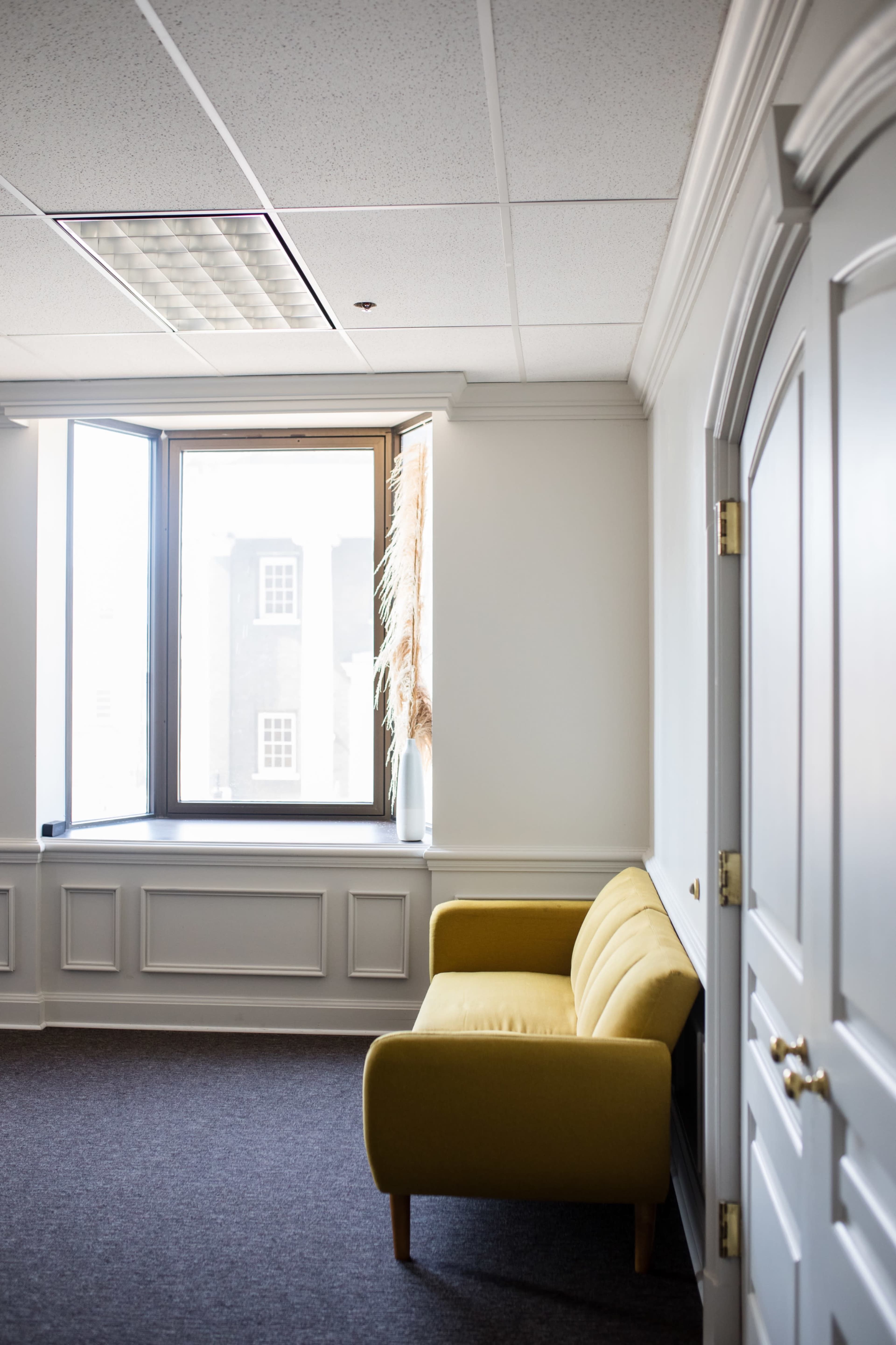 A yellow sofa is positioned beside a window in a well-lit office with light-colored walls and dark carpet.