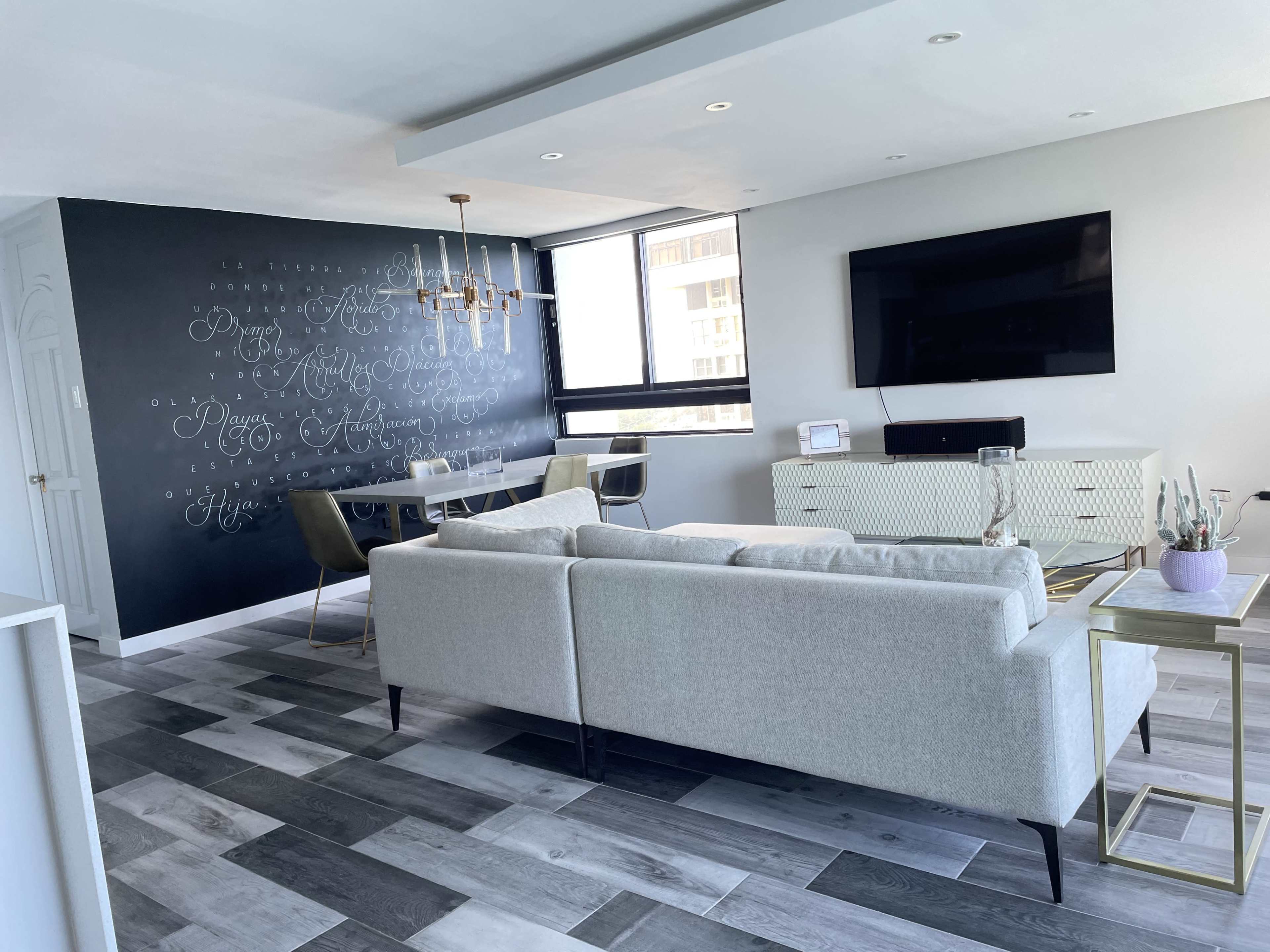 The image shows a modern living room featuring a light gray sofa, a wall decorated with handwritten text, a large television, and a dining table with chairs.