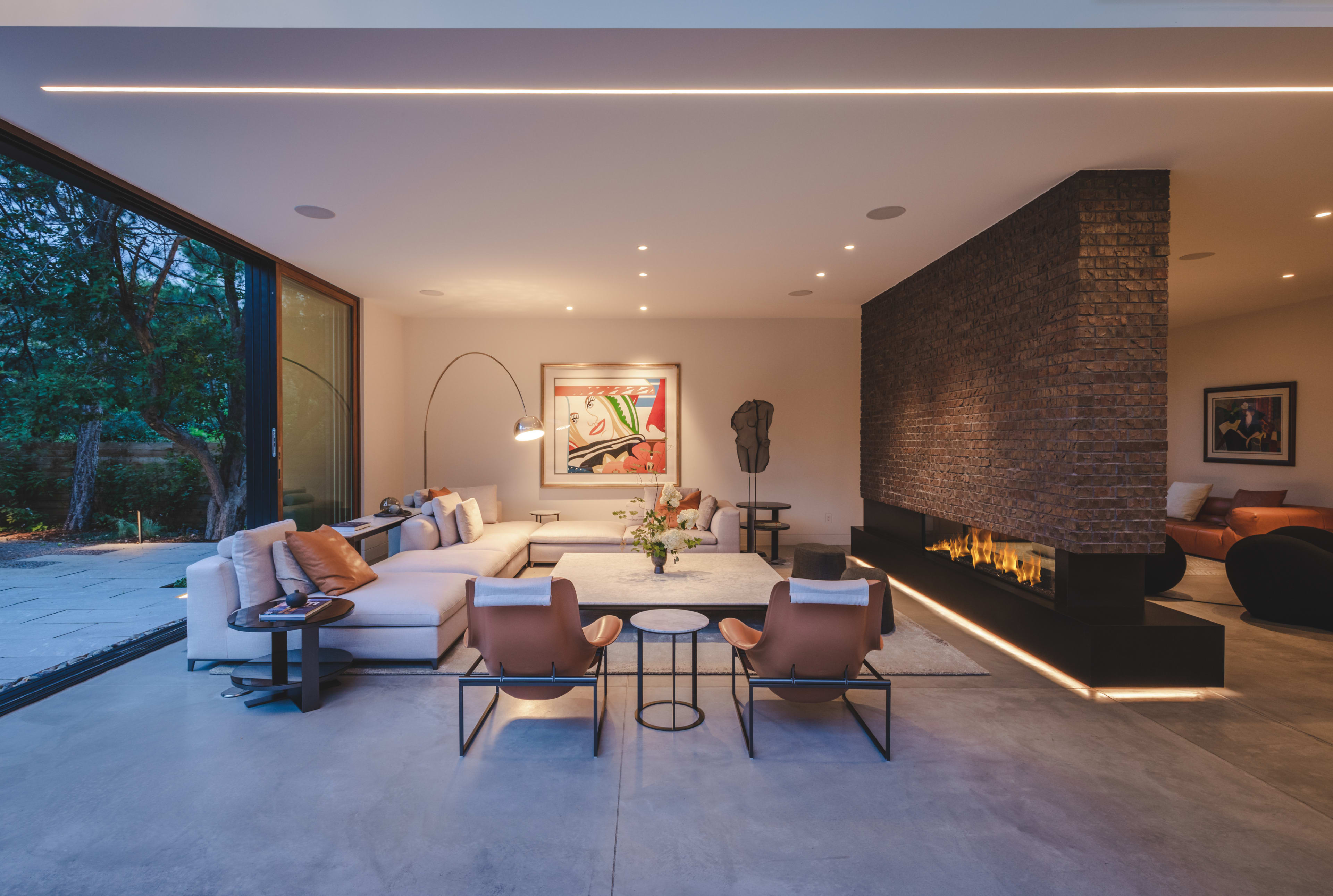 The image shows a modern living room with large windows, a sectional sofa, two armchairs, a coffee table, and a sleek fireplace, all set against a backdrop of a textured brick wall.