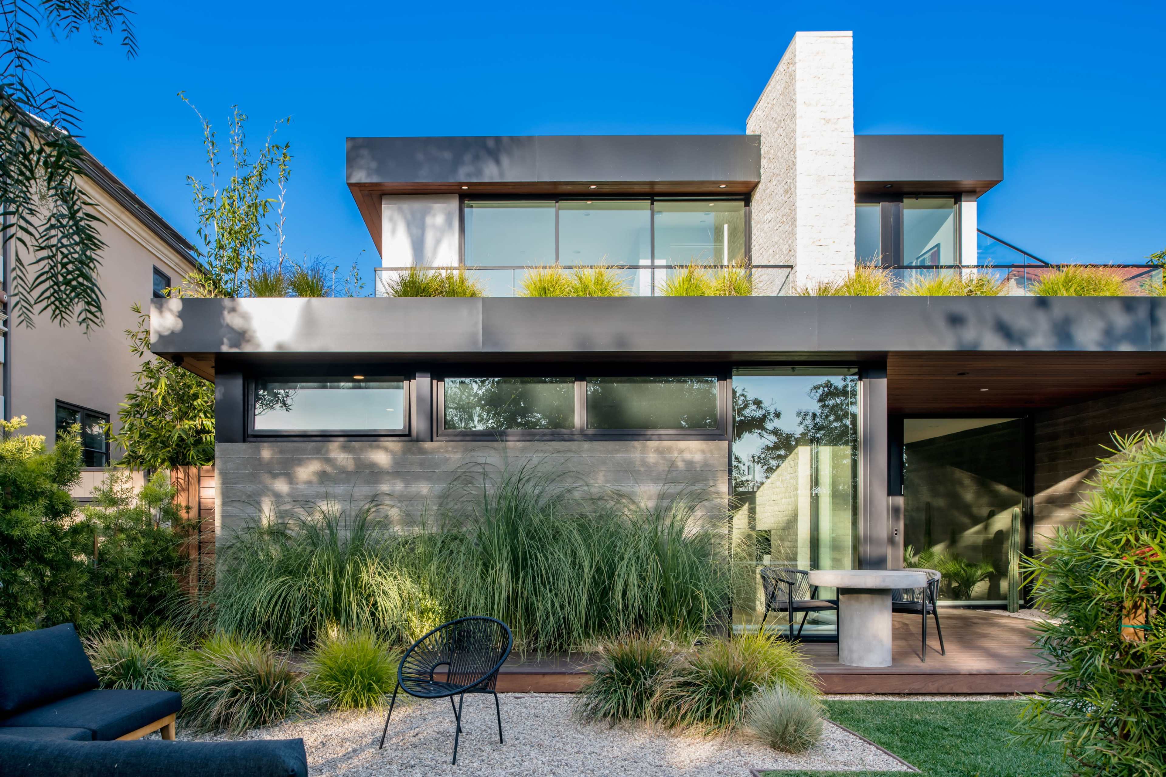 A modern two-story house with large windows, surrounded by greenery and a gravel patio area.